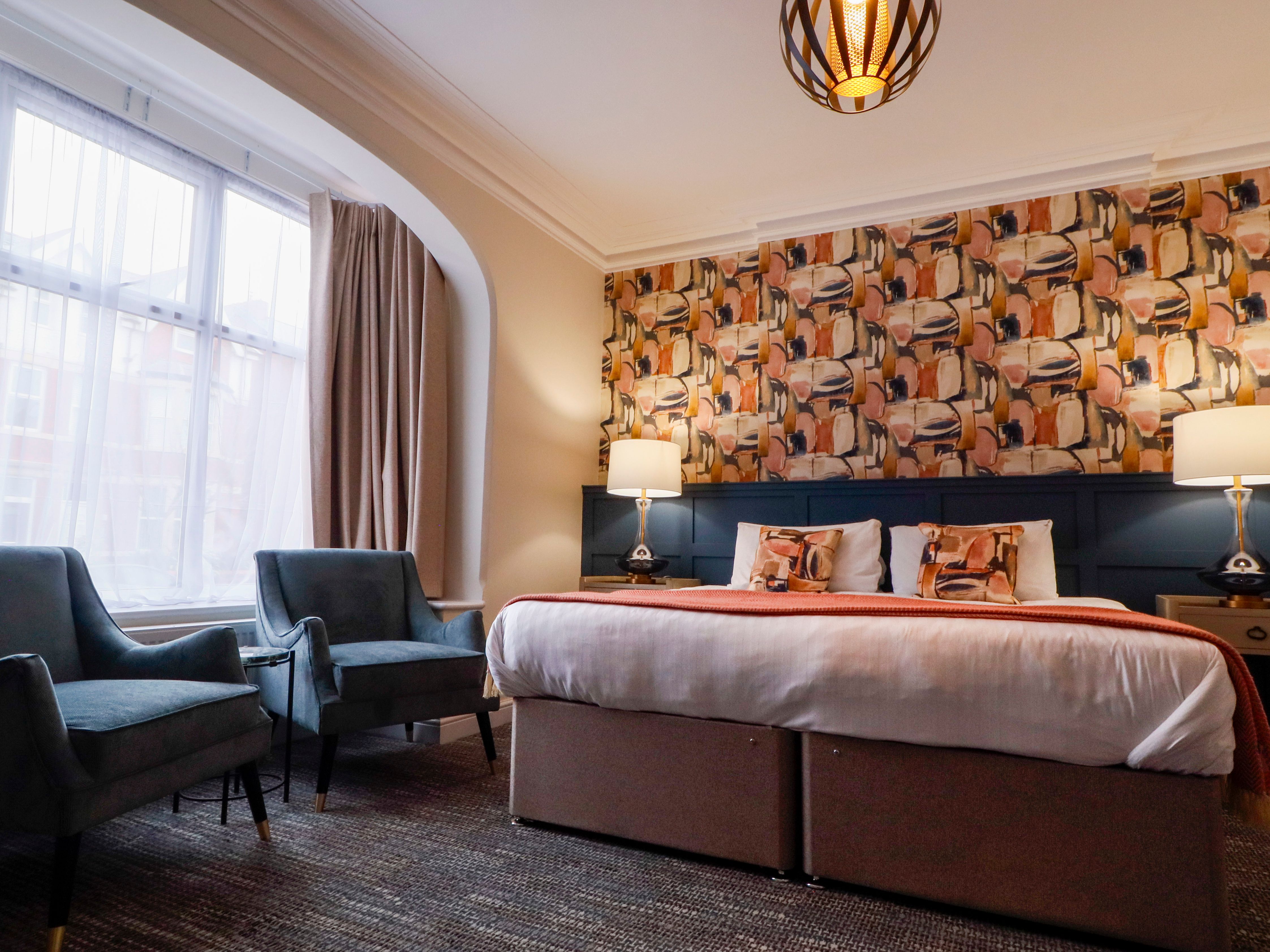 Cosy hotel bedroom with patterned feature wall, double bed, bedside lamps, and armchairs by a large arched window