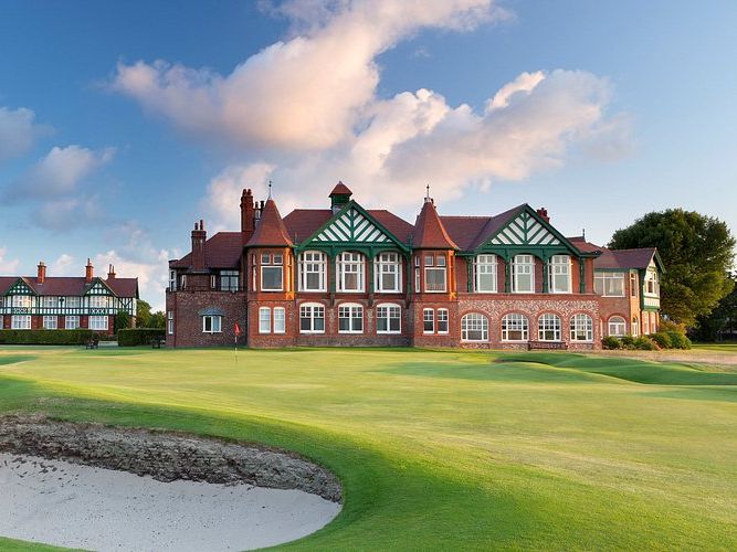 Traditional red-brick and Tudor-style clubhouse and dormy house beside a golf green with a sand bunker under a blue sky