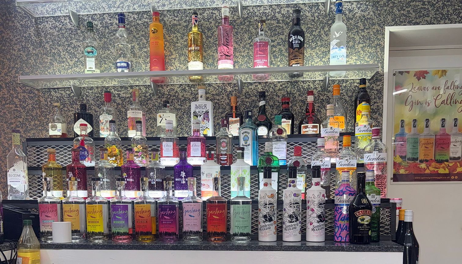 Back bar with shelves of spirits and a glass-fronted drinks fridge stocked with bottled beers and soft drinks