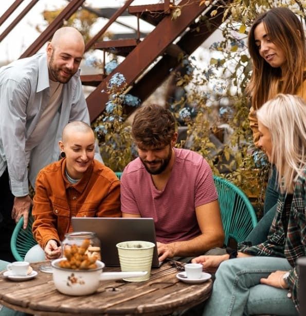 Group of six people sitting around a table outdoors, looking at a laptop and interacting with each other.