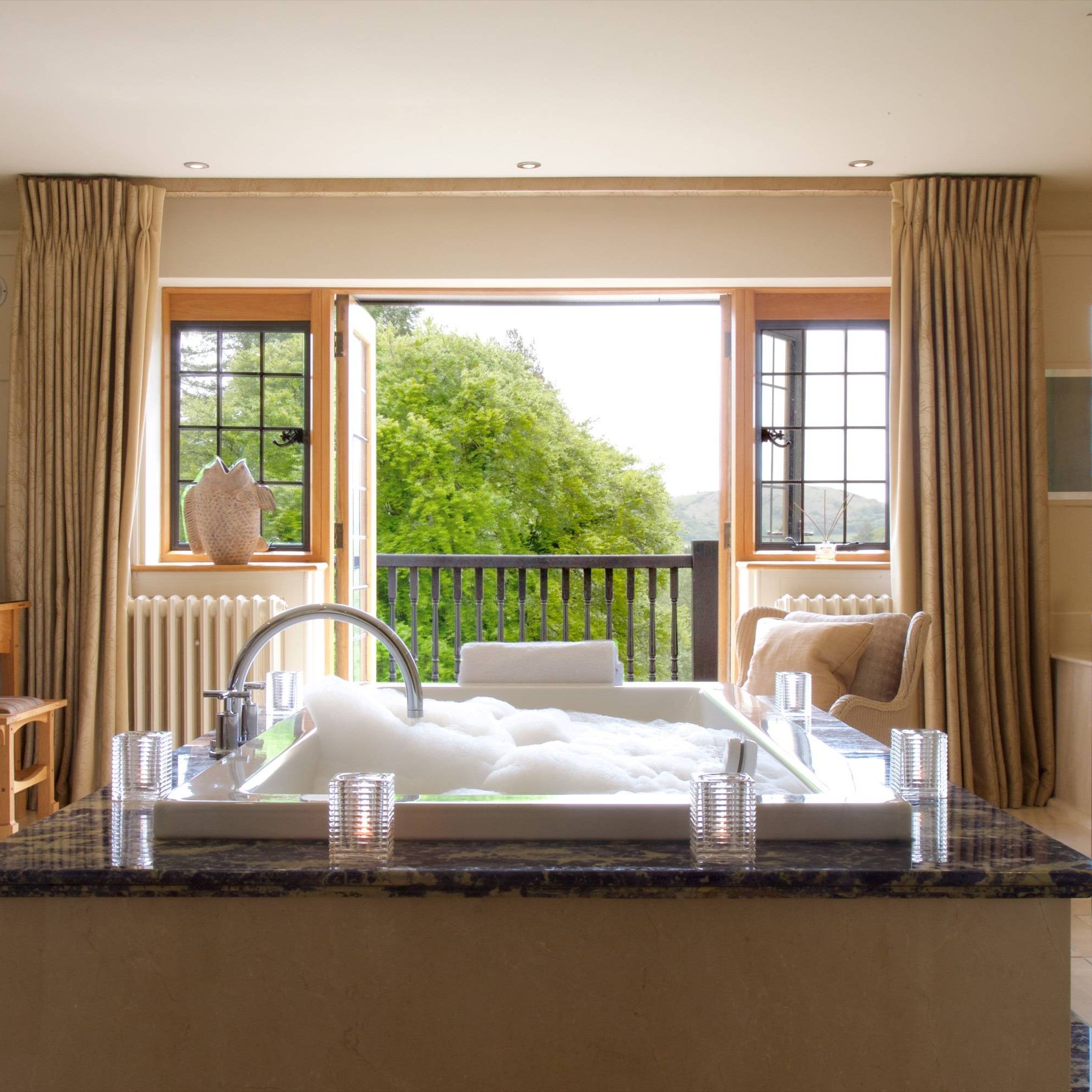 Luxurious bathroom with a bubble-filled bathtub, open windows with a view of lush greenery, and cozy seating area.