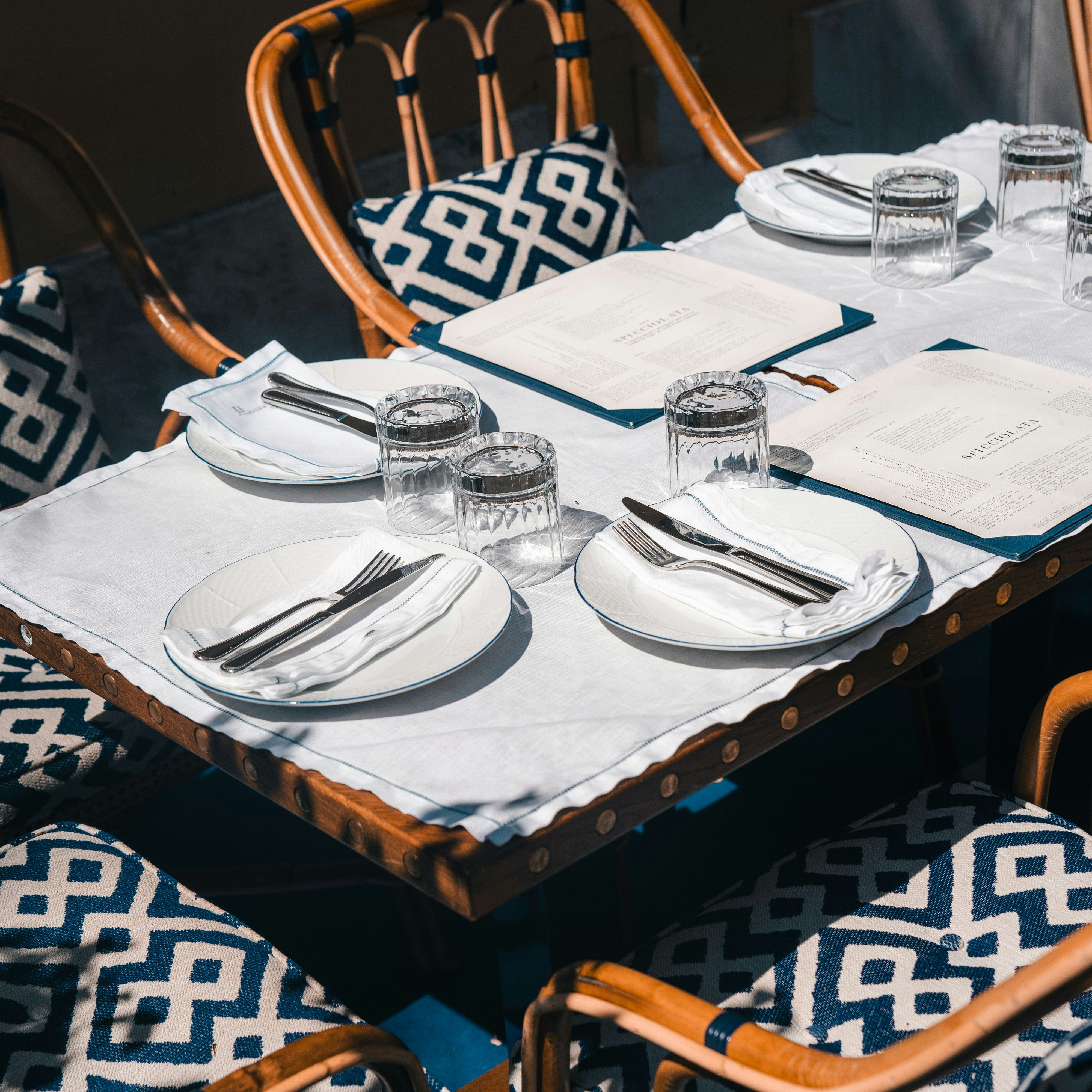 Outdoor restaurant table set with plates, cutlery, glasses, menus, and patterned cushioned chairs