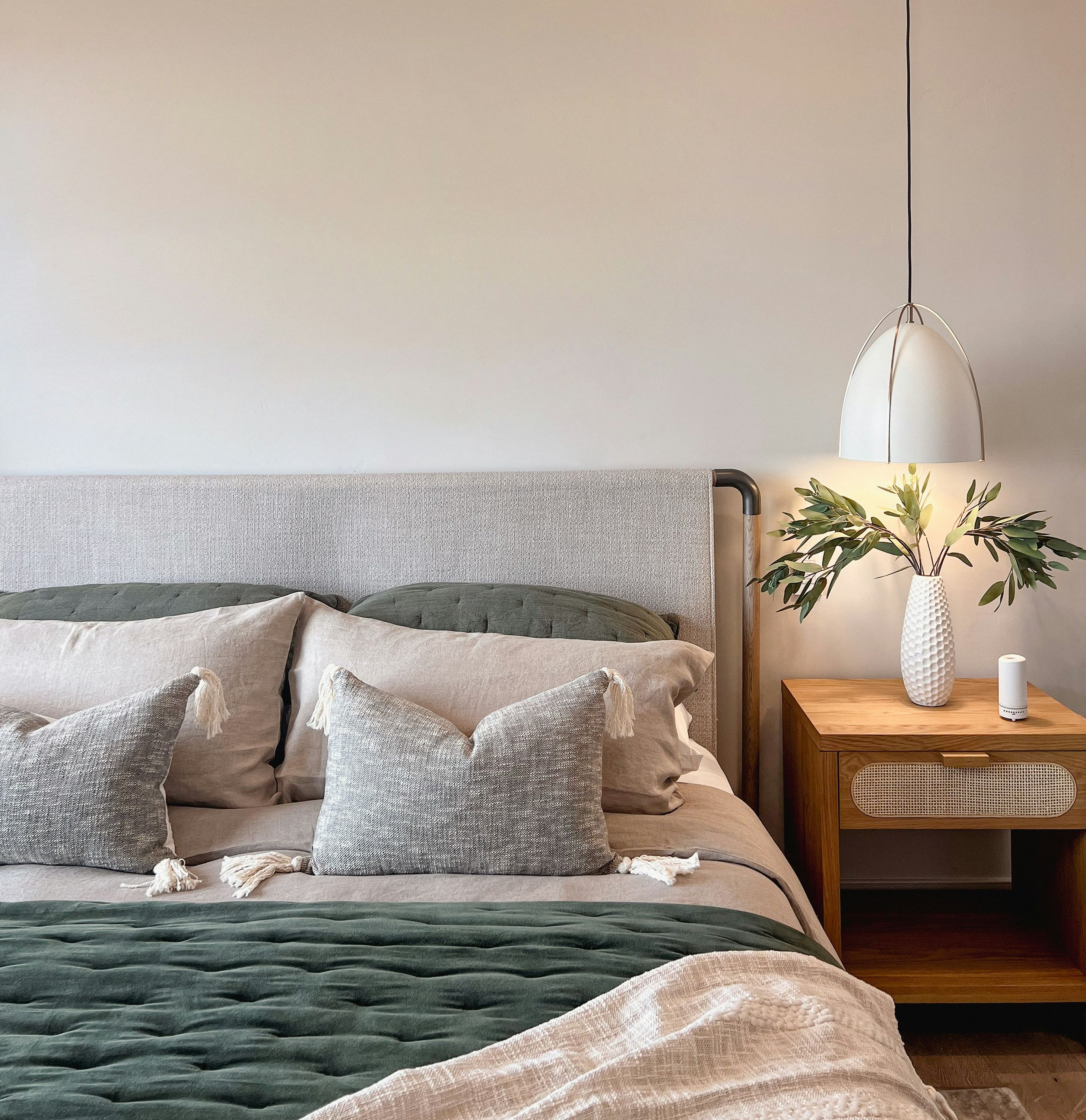 Modern bedroom with a grey headboard, neutral bedding, decorative pillows, a wooden nightstand, a vase with greenery, and a pendant lamp.