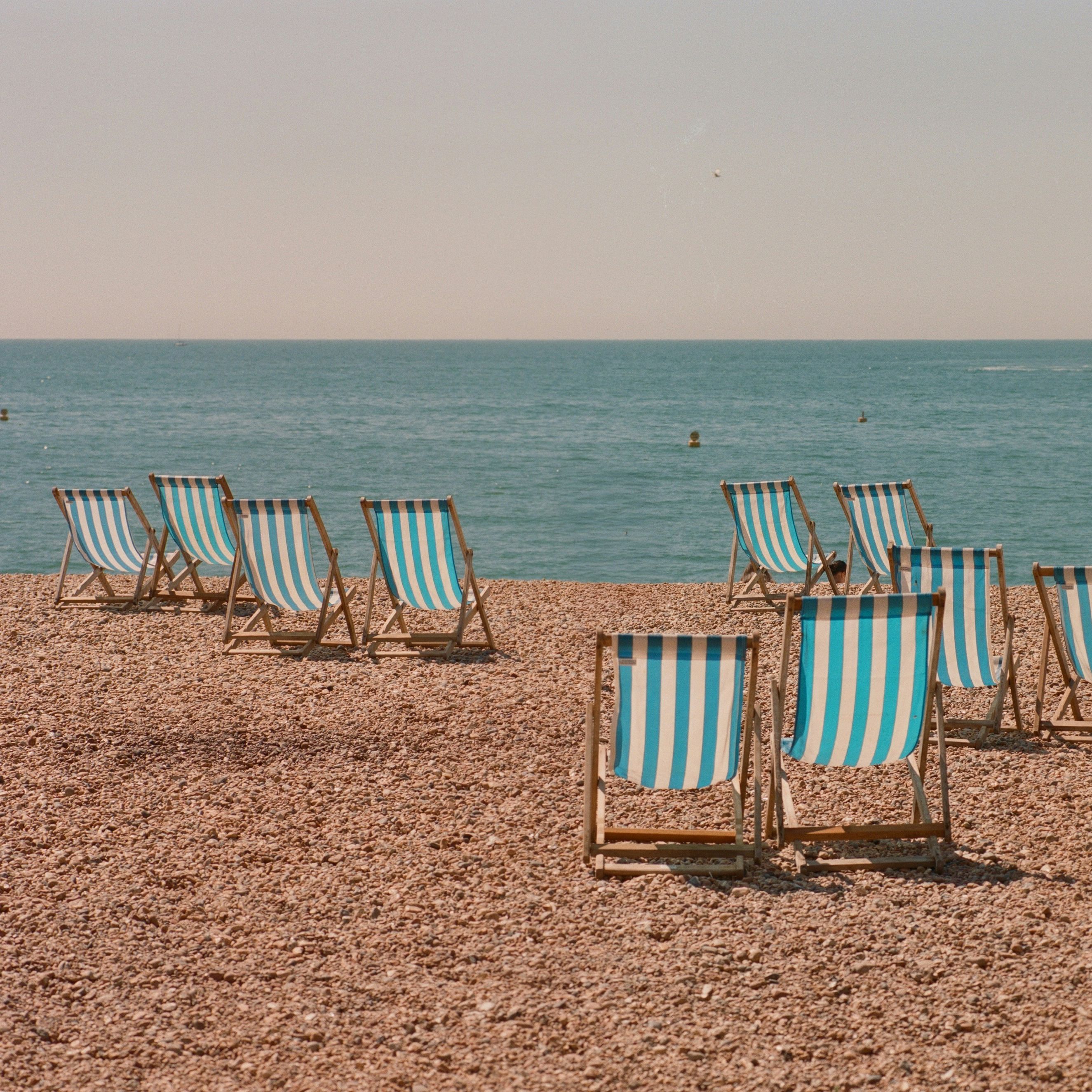 Striped deck chairs arranged on a pebbly beach facing the sea, with a 'For Hire' sign visible.