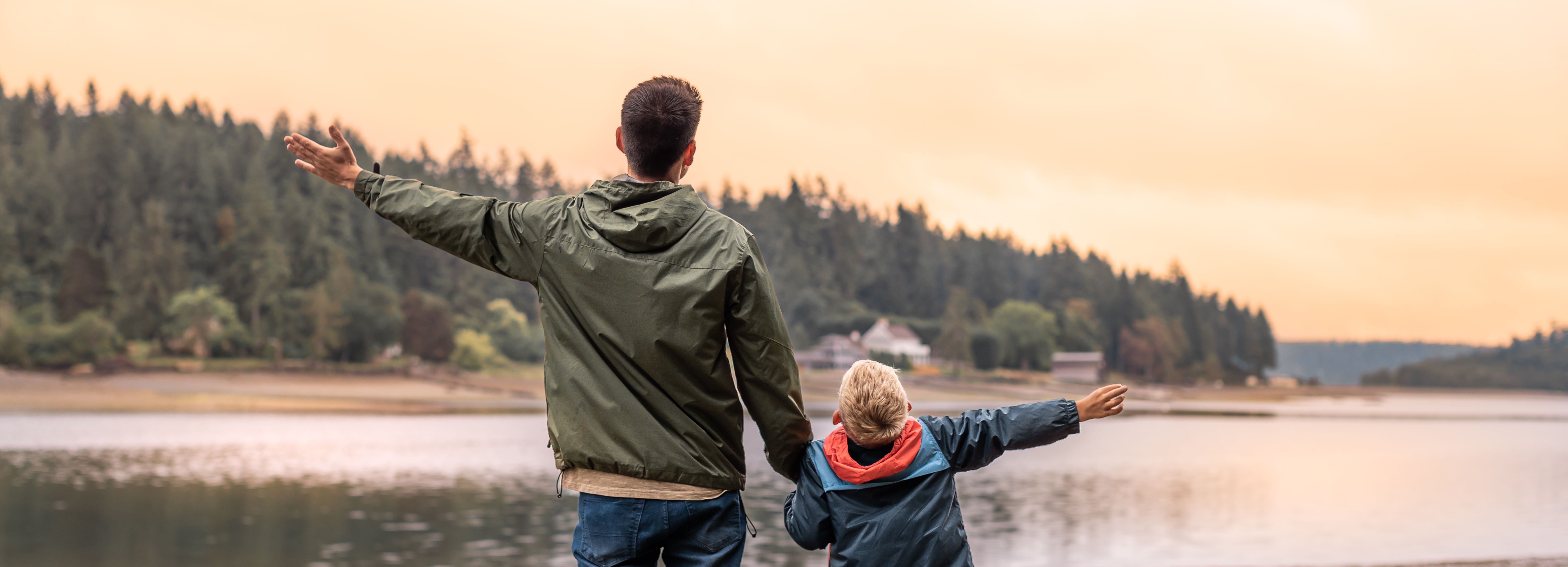 Father and son standing by a lake with arms outstretched, facing the water during sunset.