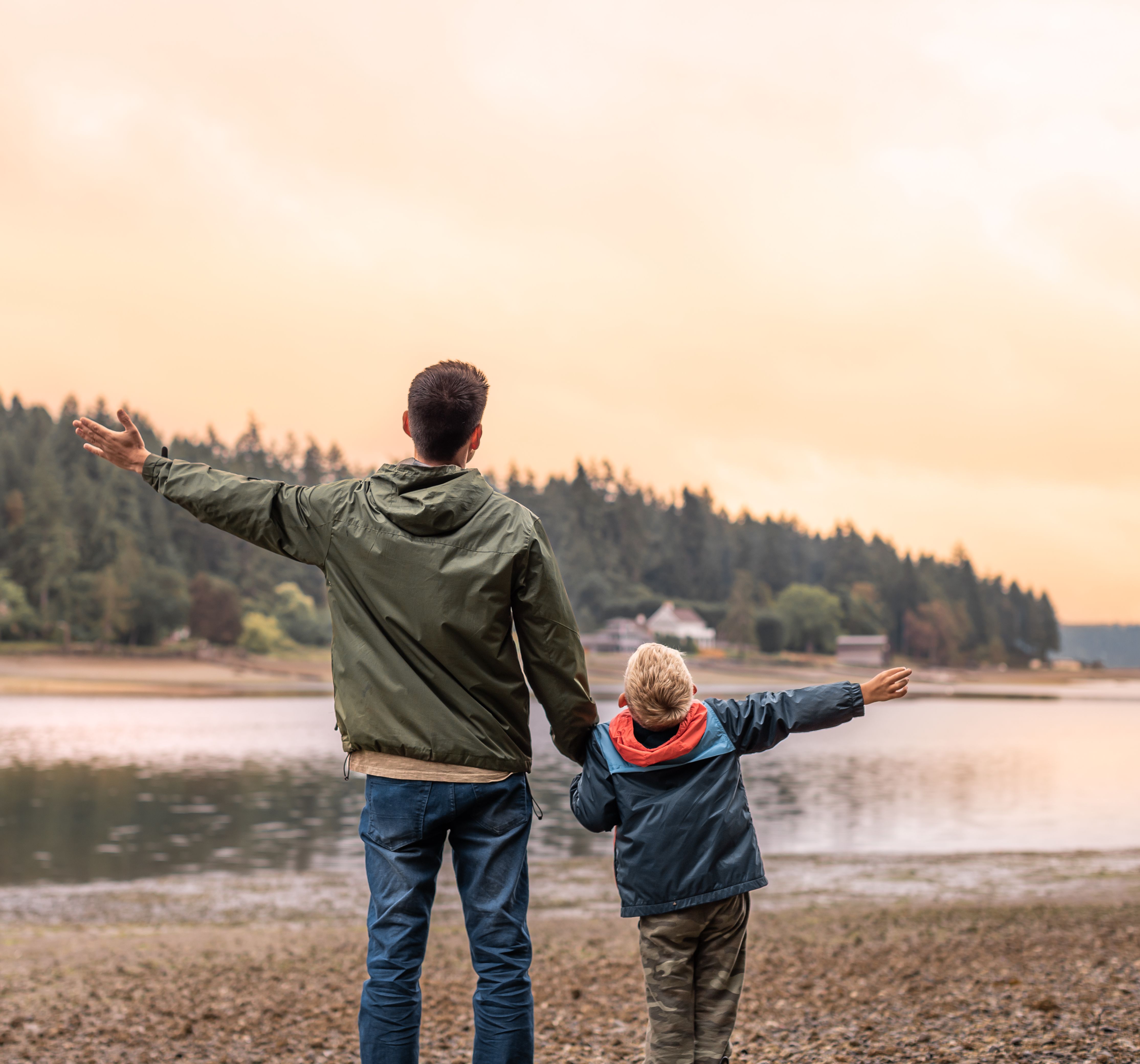Father and son standing by a lake with arms outstretched, facing the water during sunset.