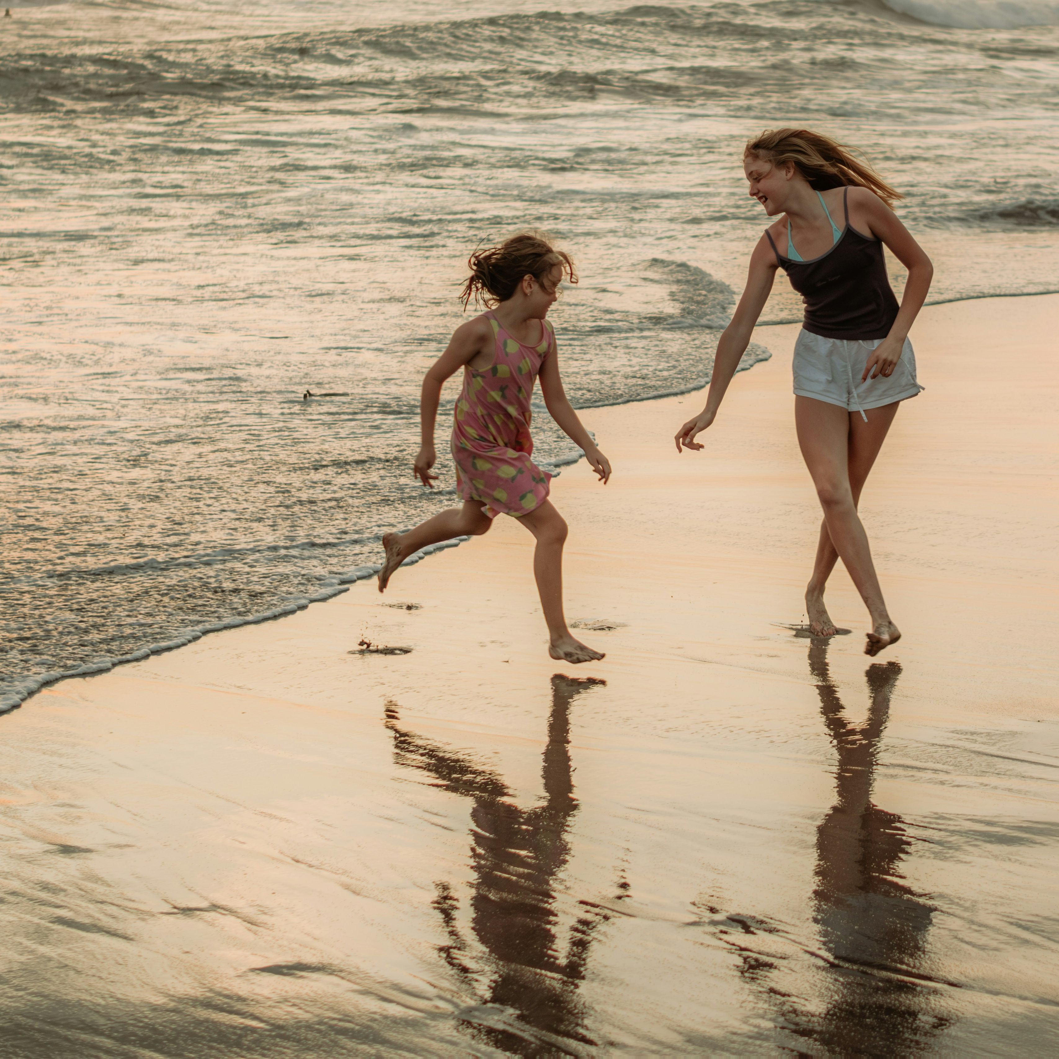Woman and girl running and playing on the beach at sunset, their reflections visible in the wet sand.