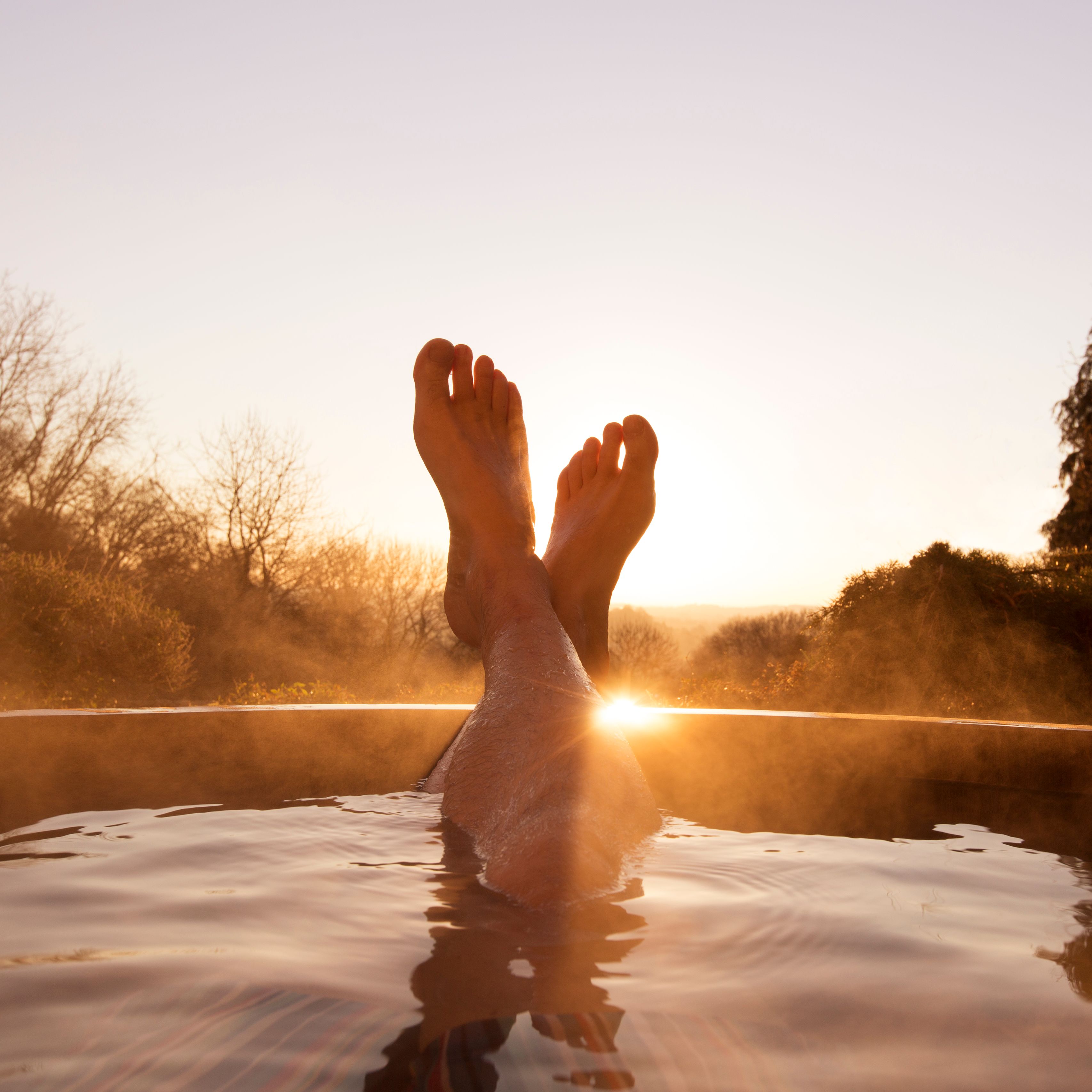 Person relaxing in an outdoor hot tub at sunset with legs crossed and steam rising from the water.