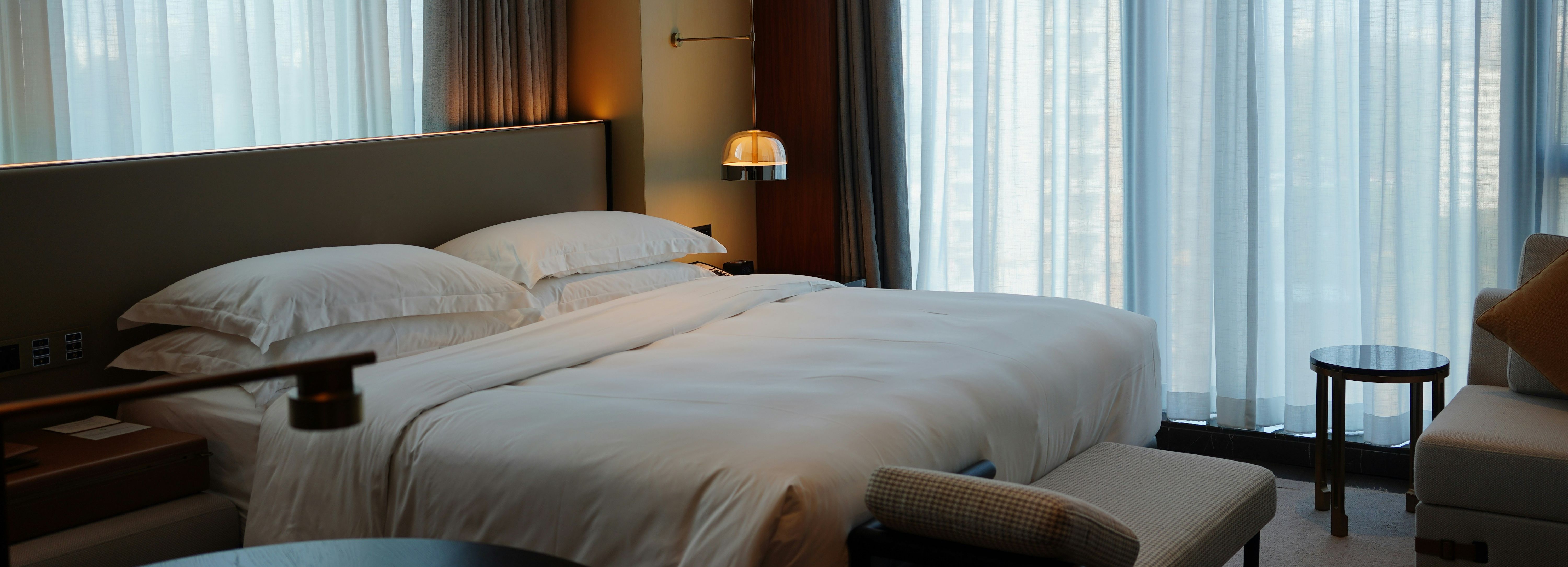 Modern hotel room interior with large bed, white linens, and floor-to-ceiling windows with sheer curtains