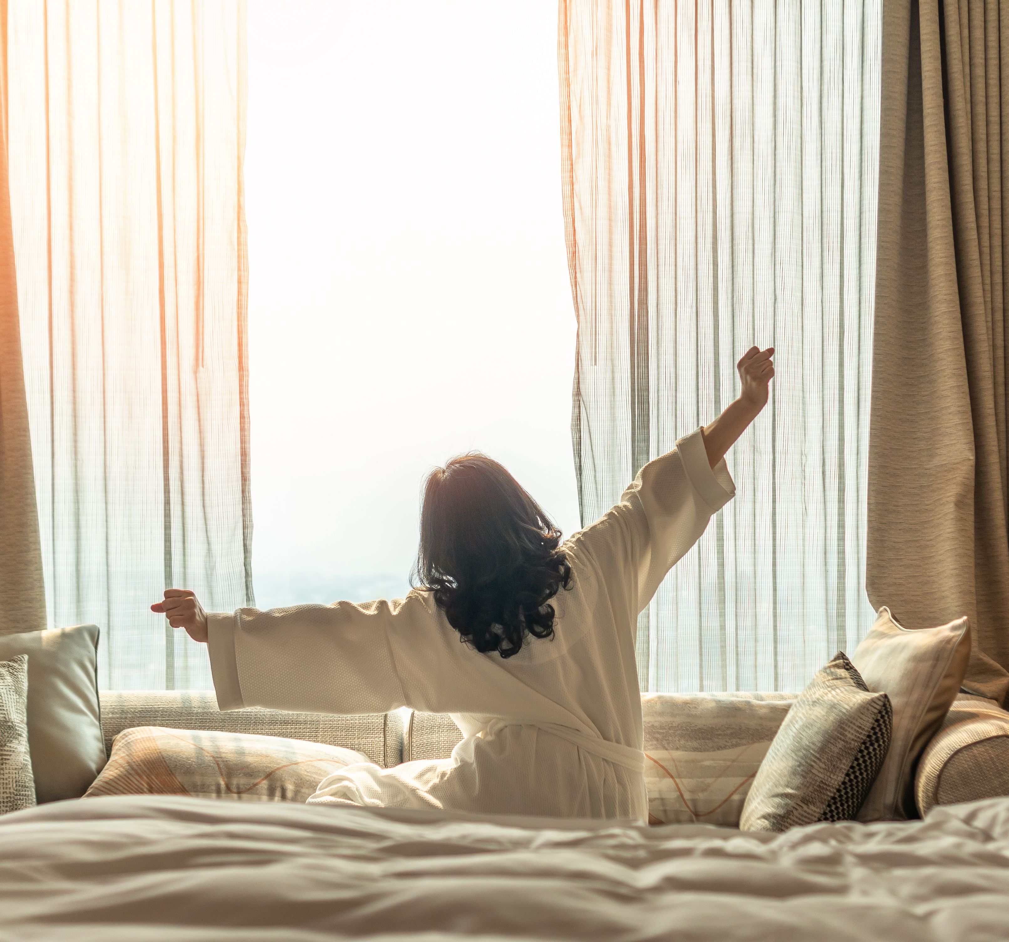 Woman stretching in bed with morning sunlight streaming through curtains