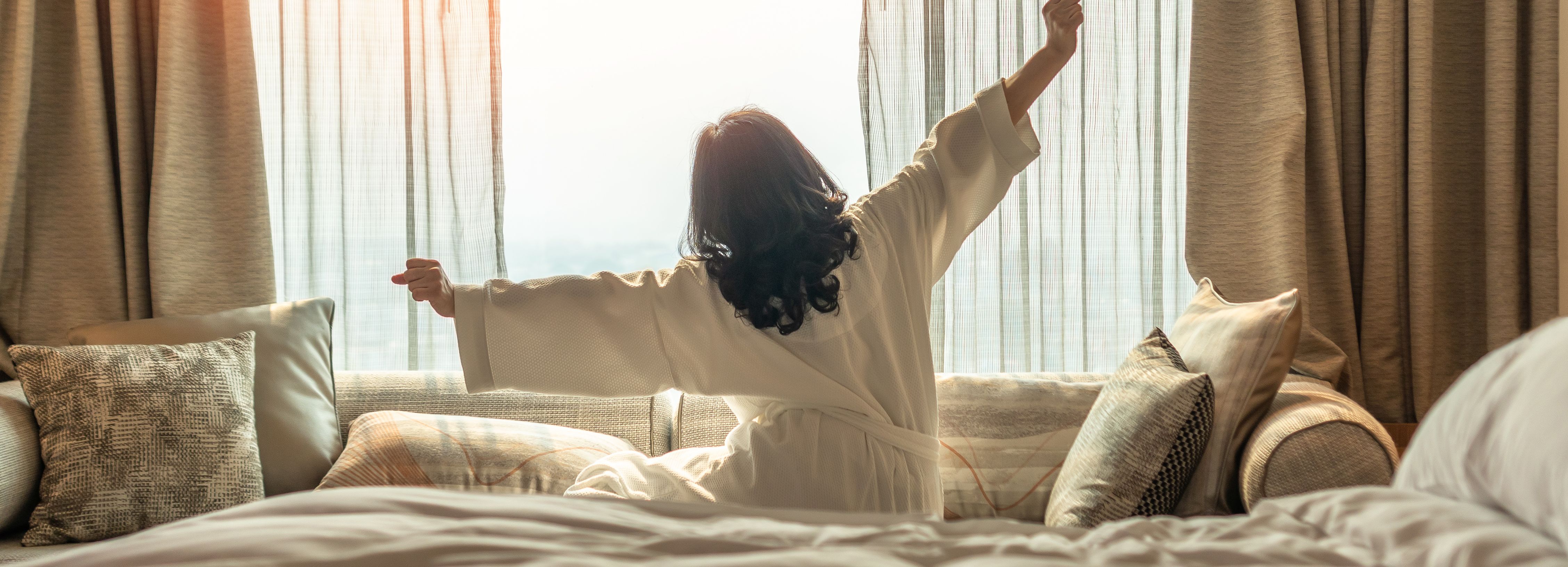 Woman stretching in bed with morning sunlight streaming through curtains