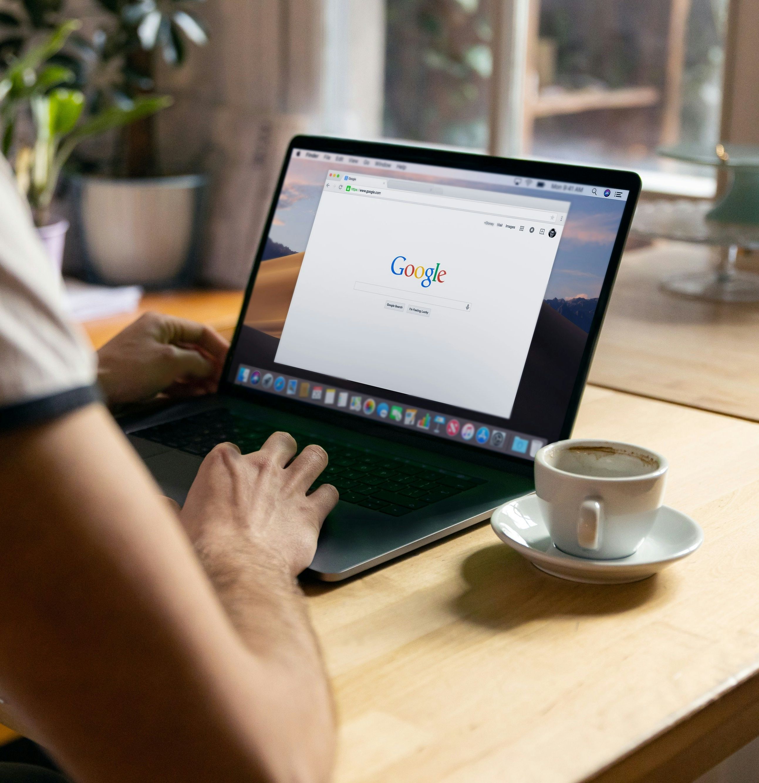 Person using a laptop with Google homepage open on the screen, sitting at a wooden table with a cup of coffee.