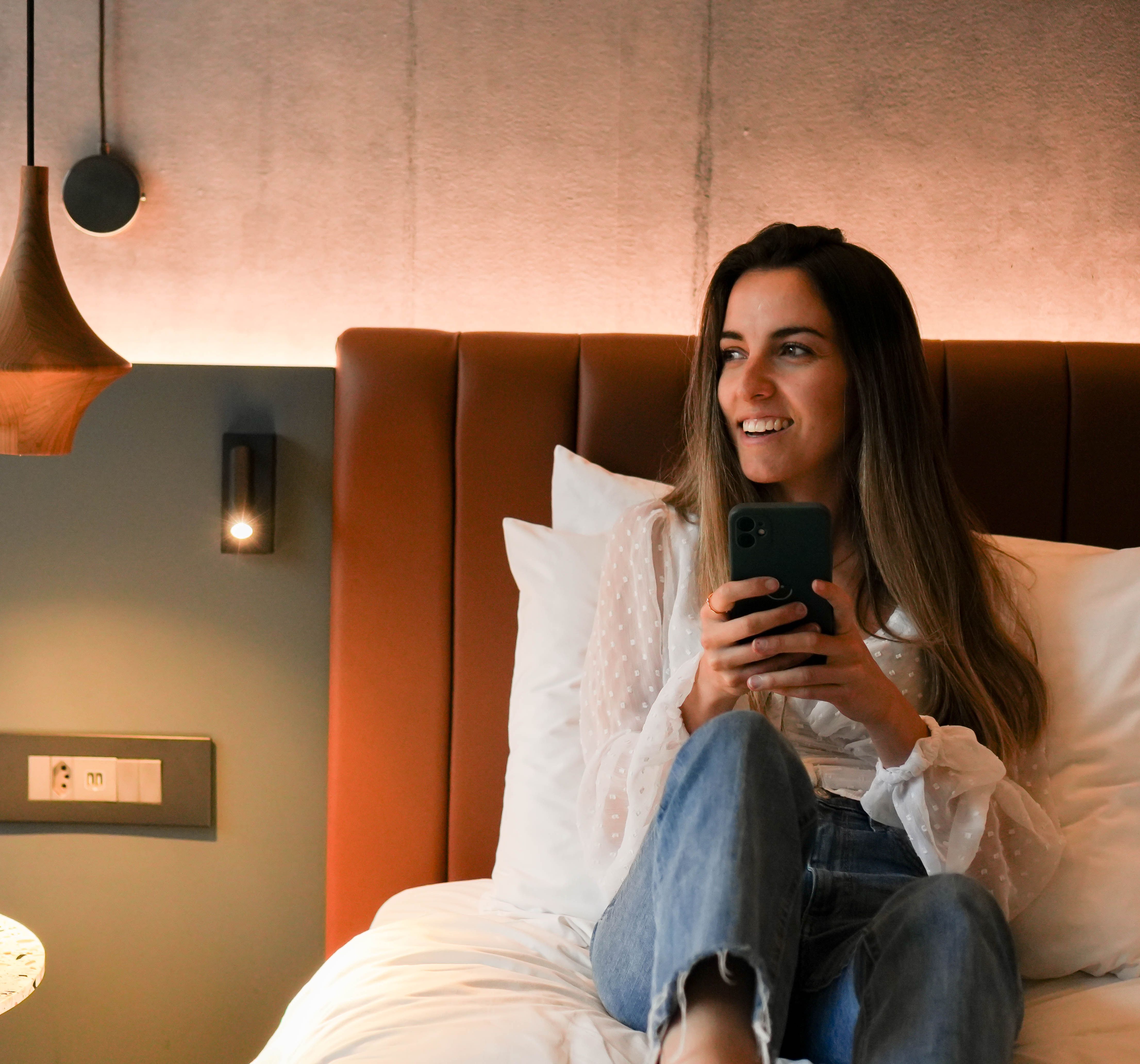 Woman sitting on bed holding a phone and smiling in a modern cozy room