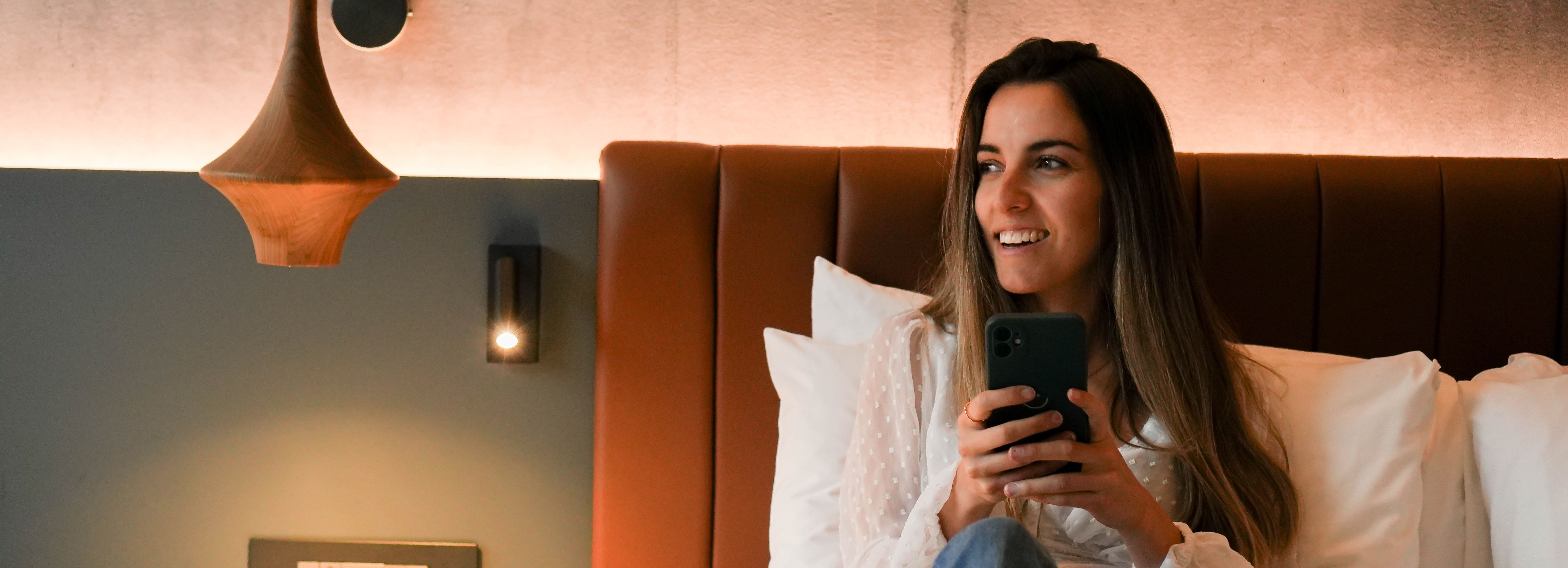 Woman sitting on bed holding a phone and smiling in a modern cozy room