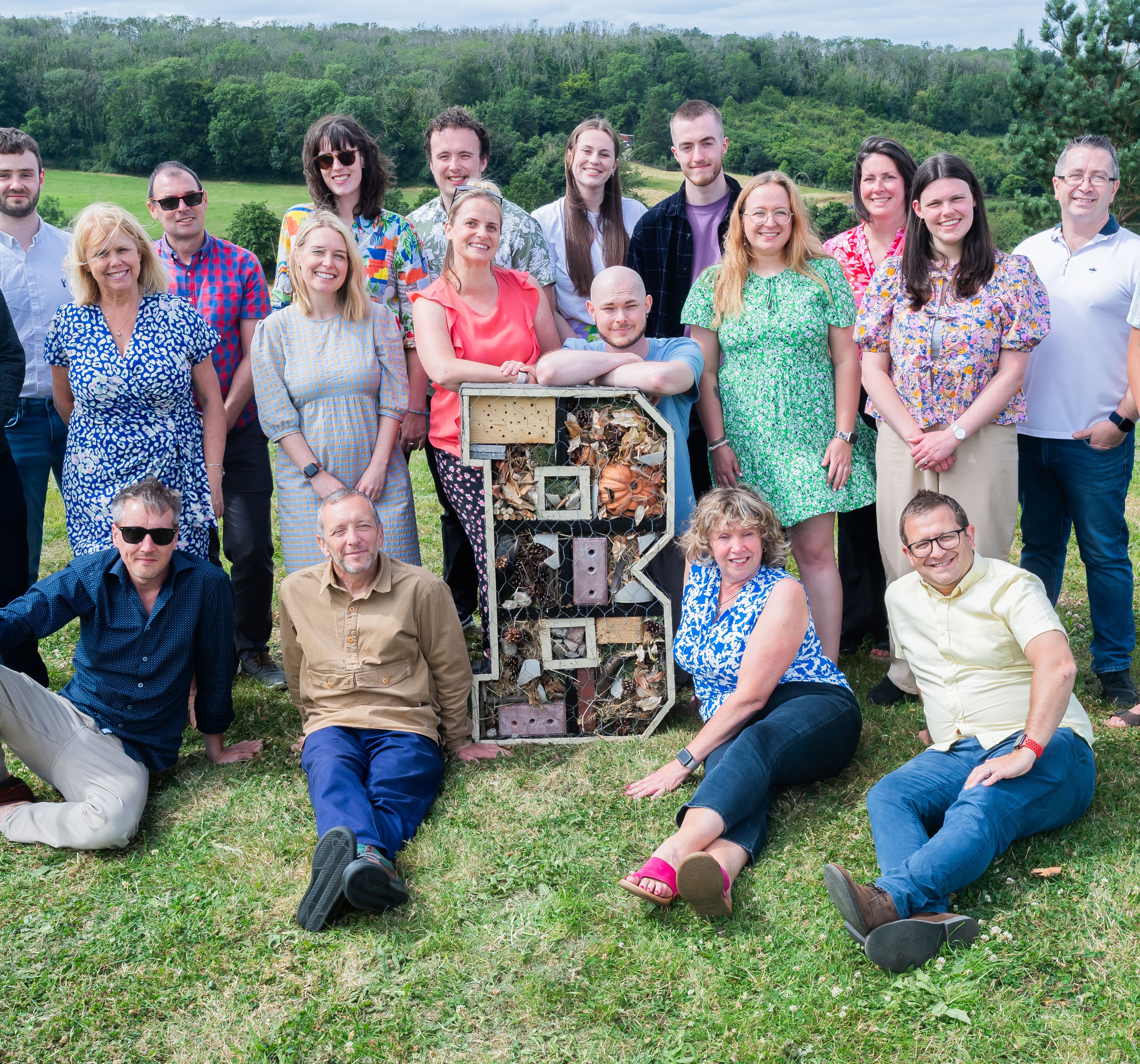 A group of people posing outdoors on grass with trees in the background, gathered around a large letter 'B' structure filled with natural materials.