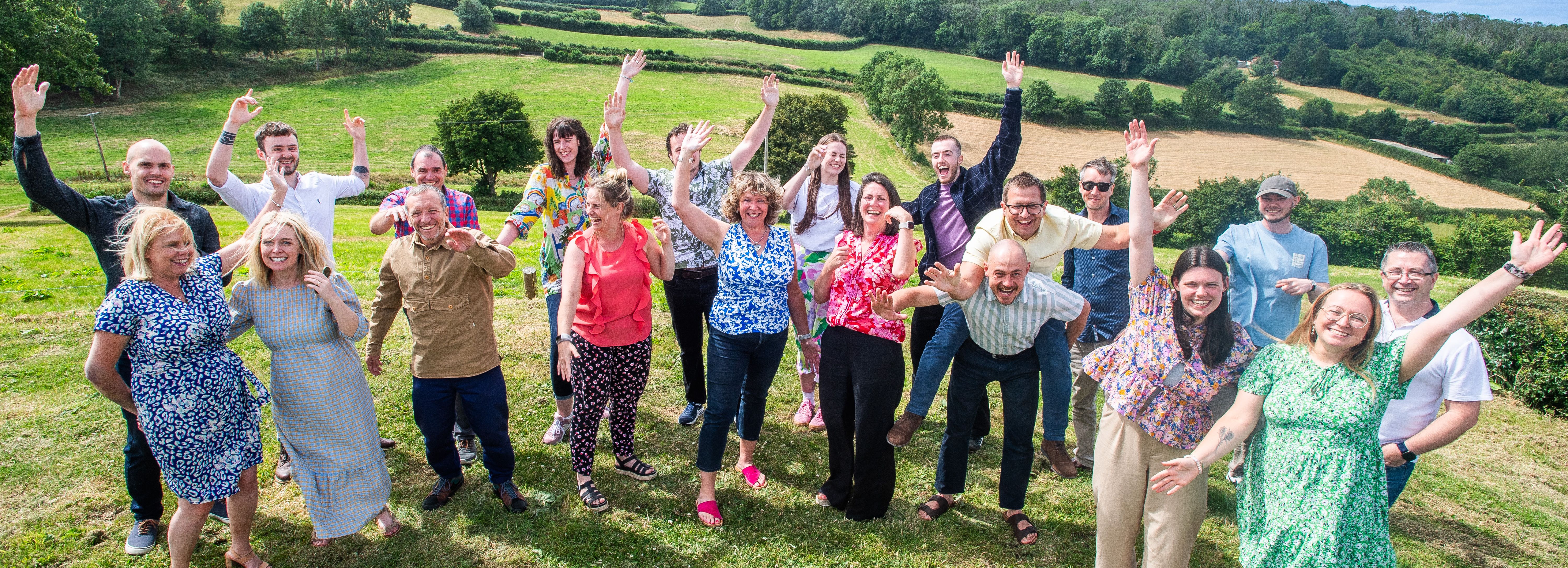 Group of people posing and smiling with arms raised outdoors on grassy hill with scenic countryside in the background.