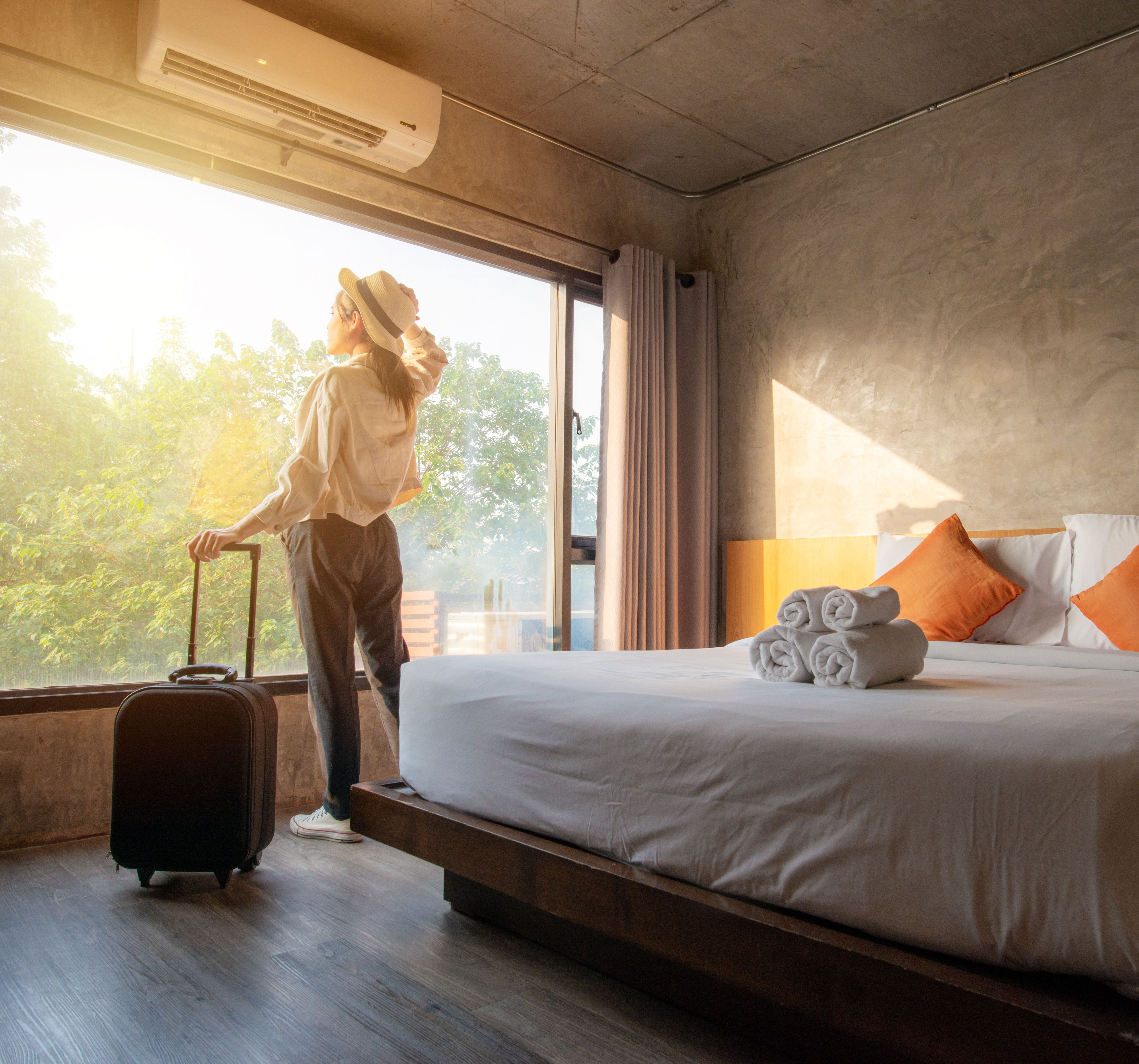 Woman with suitcase looking out window in a modern hotel room with a large bed and sunlight streaming in.