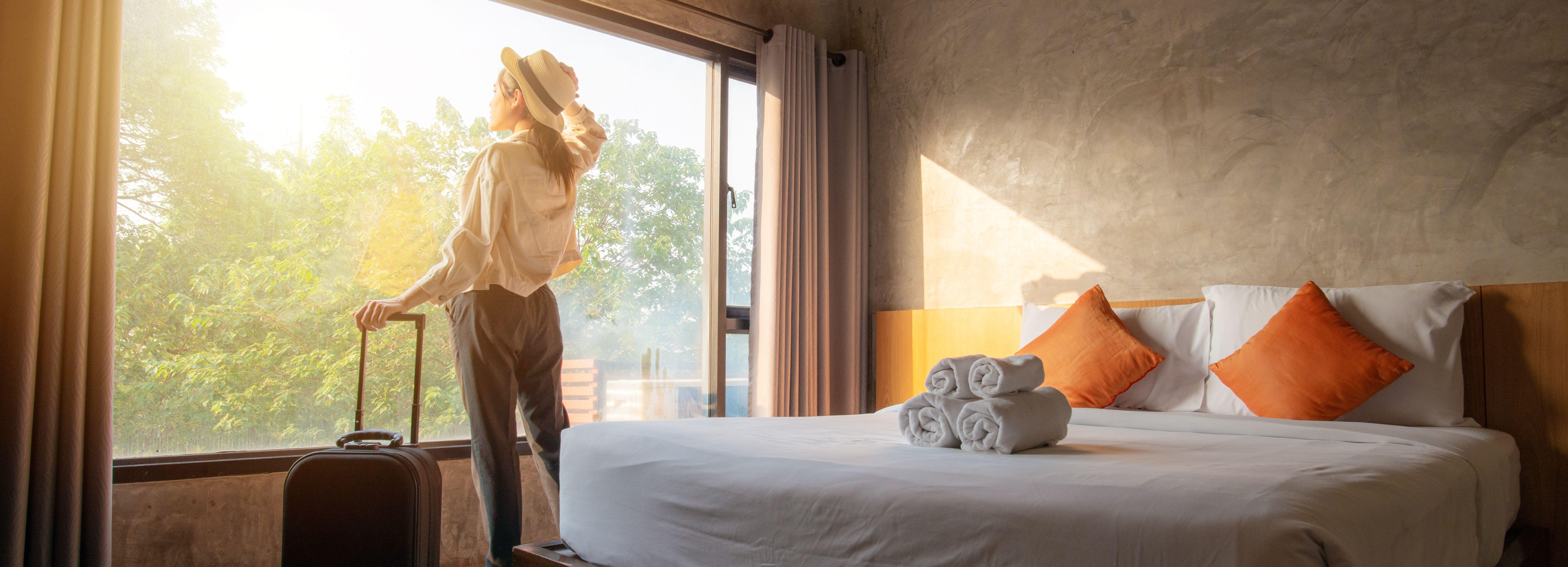 Woman with suitcase looking out window in a modern hotel room with a large bed and sunlight streaming in.