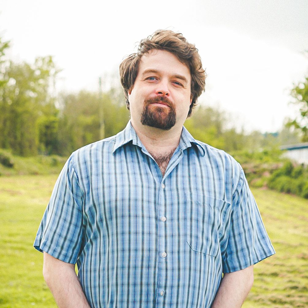 Man with brown hair and beard standing outdoors on a grassy field, wearing a blue plaid shirt