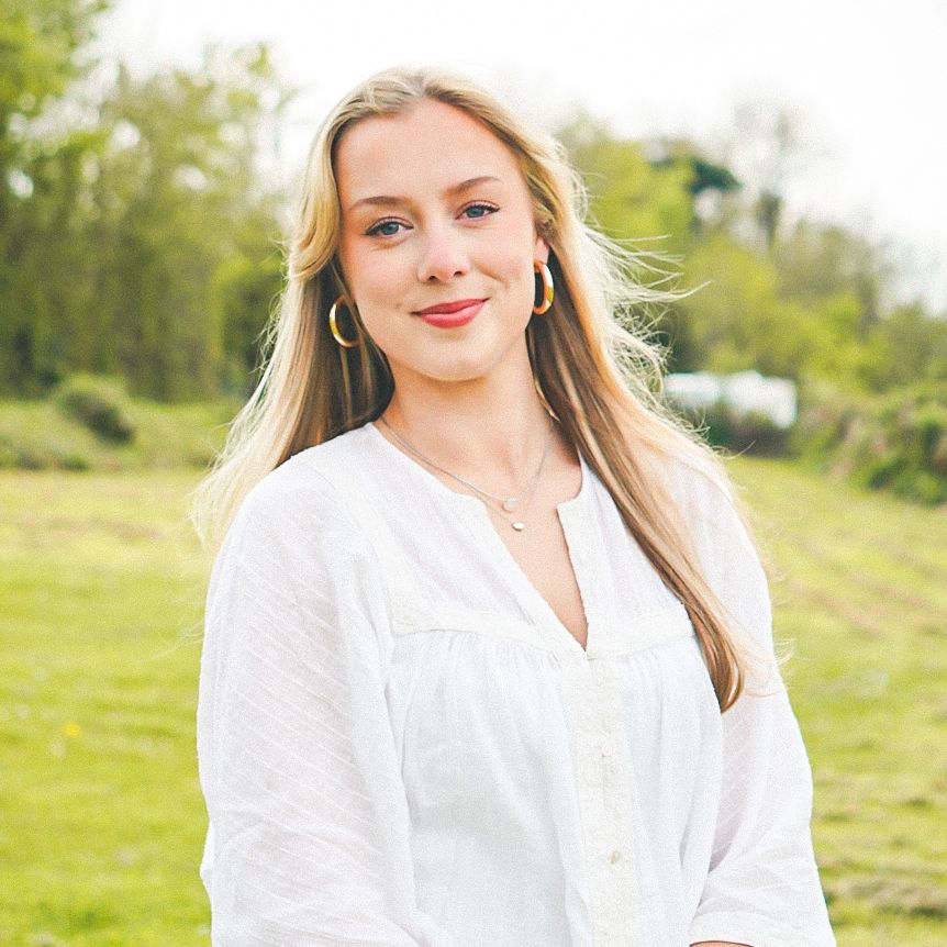Young woman with long blonde hair standing in a grassy field, wearing a white blouse and blue jeans