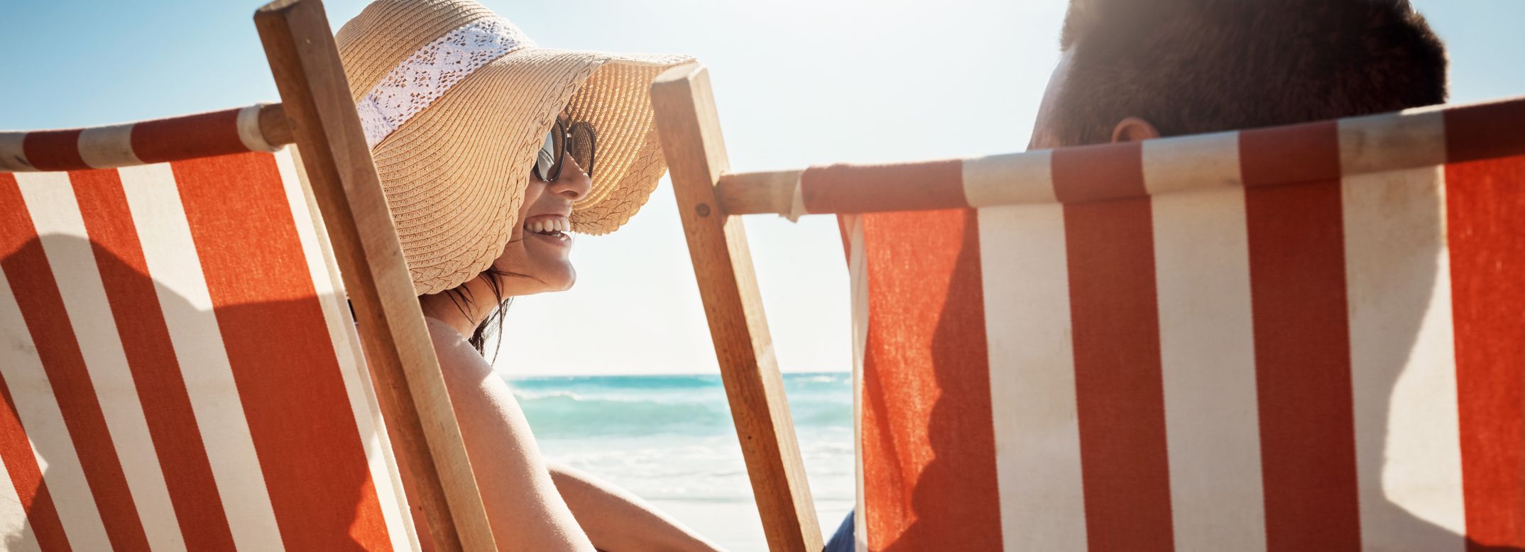 Woman wearing a sunhat and sunglasses sitting on a striped deck chair at the beach, smiling and facing a companion.