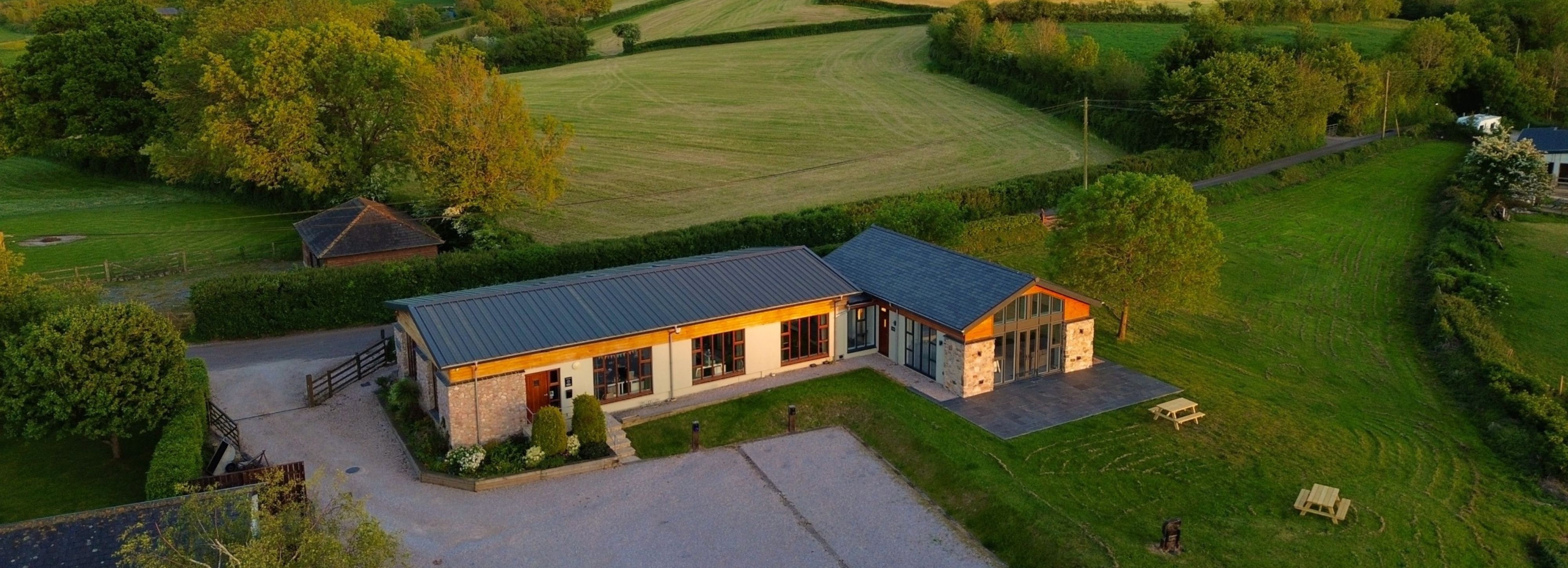Modern country house surrounded by green fields and trees, viewed from above at sunset.