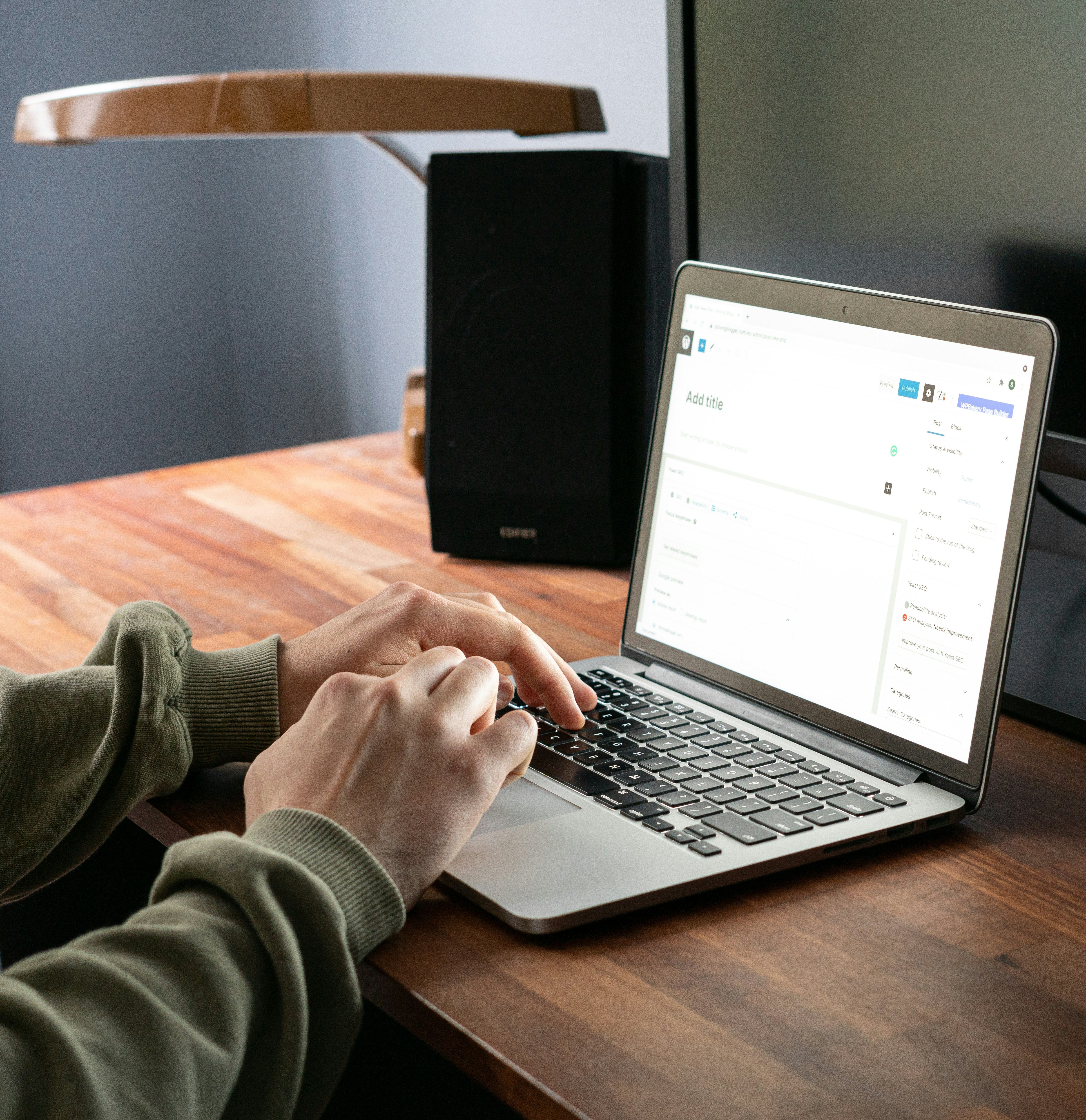 Person typing on a laptop at a wooden desk with a monitor and speaker