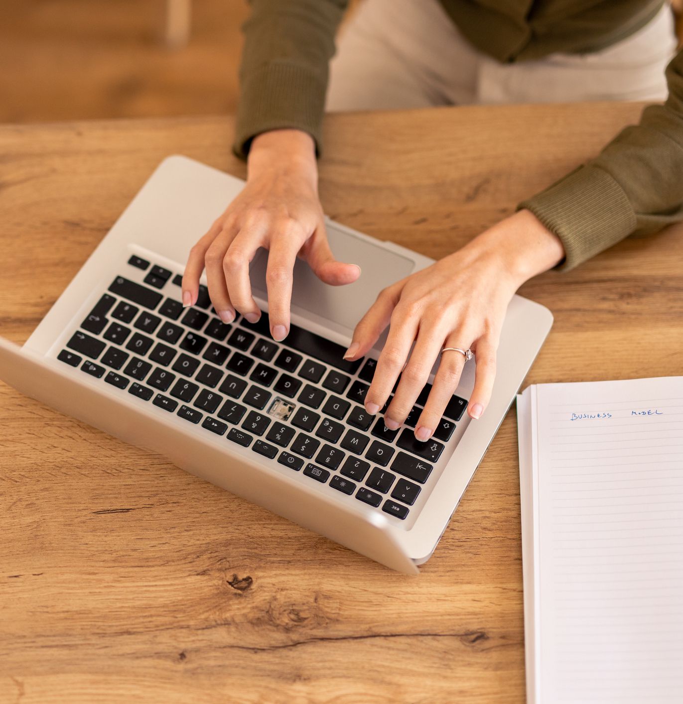Person typing on a laptop with a notebook, smartphone, and cup on a wooden desk.