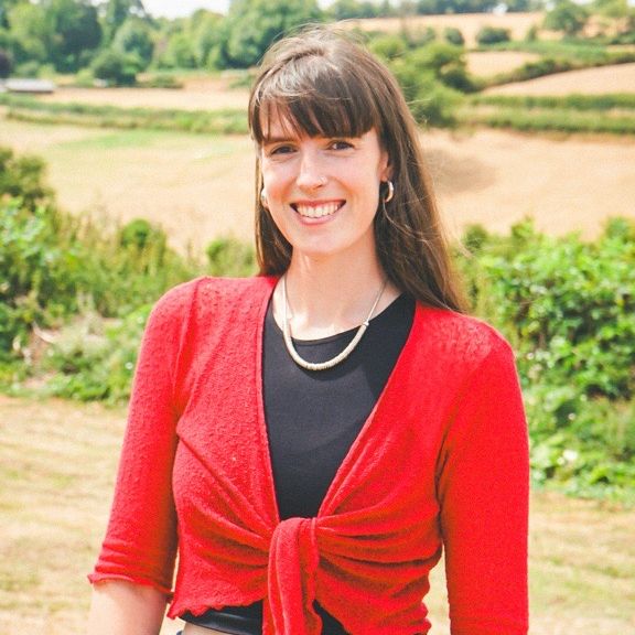 Woman smiling outdoors, wearing a red top and blue jeans with a countryside background.