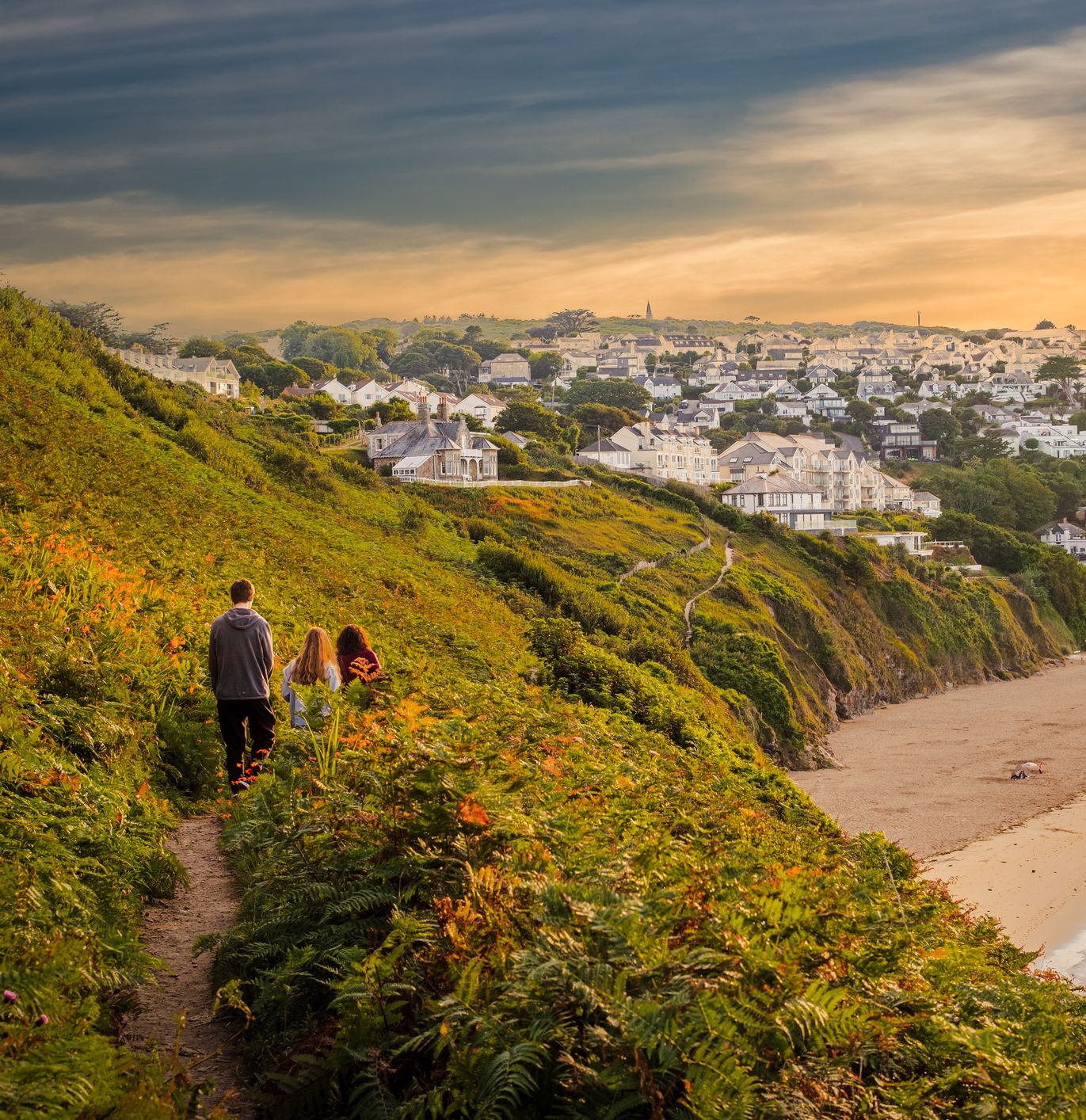 Three people walking along a coastal cliff path above a sandy beach, with a seaside town in the background at sunset.