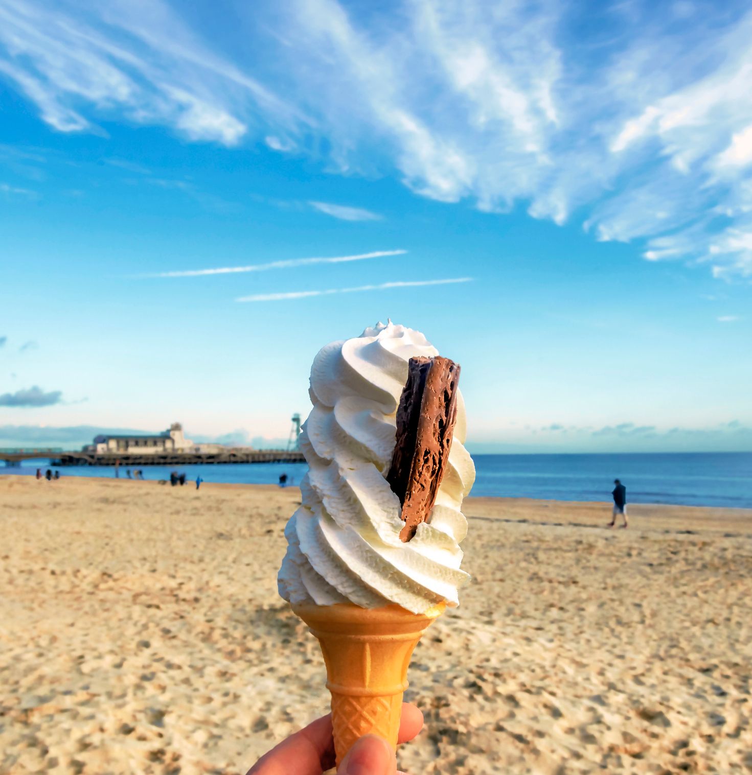 Hand holding a soft serve ice cream cone with a chocolate flake on a sandy beach with the sea and pier in the background