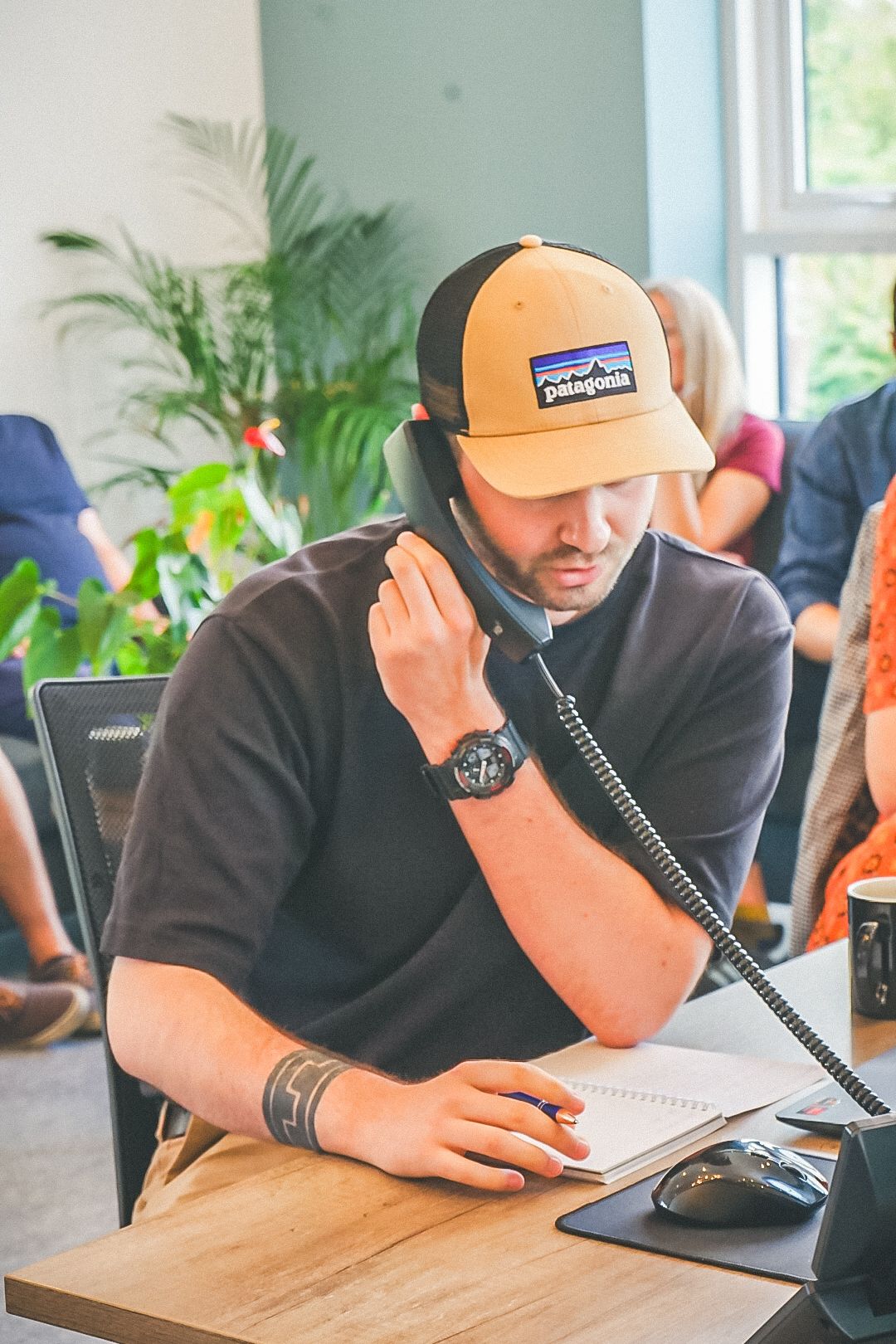 Man wearing a Patagonia cap talking on a desk phone and taking notes at an office desk