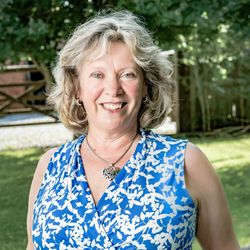 Smiling woman with curly blonde hair wearing a blue and white floral sleeveless top standing outdoors.