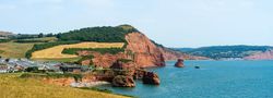 Scenic view of red sandstone cliffs by the sea with green fields and coastal buildings under a blue sky.