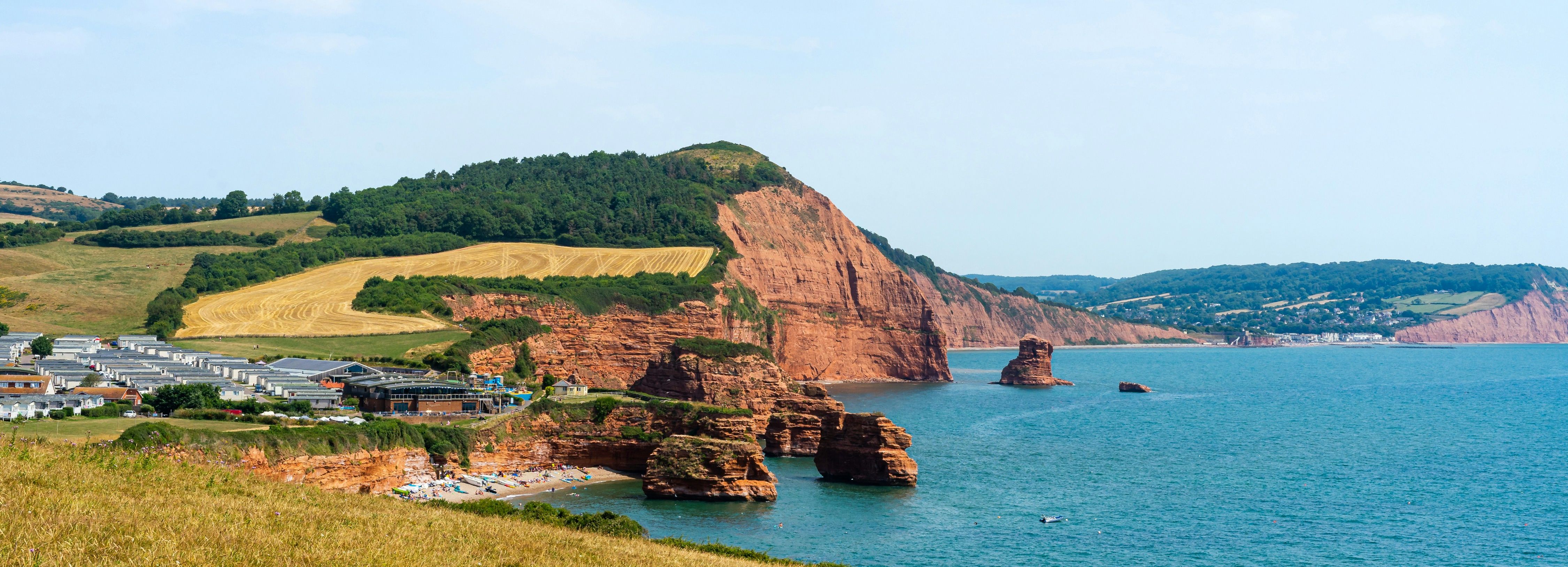 Scenic view of red sandstone cliffs by the sea with green fields and coastal buildings under a blue sky.