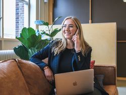 Woman sitting on a sofa with a laptop, smiling and talking on the phone in a modern office setting