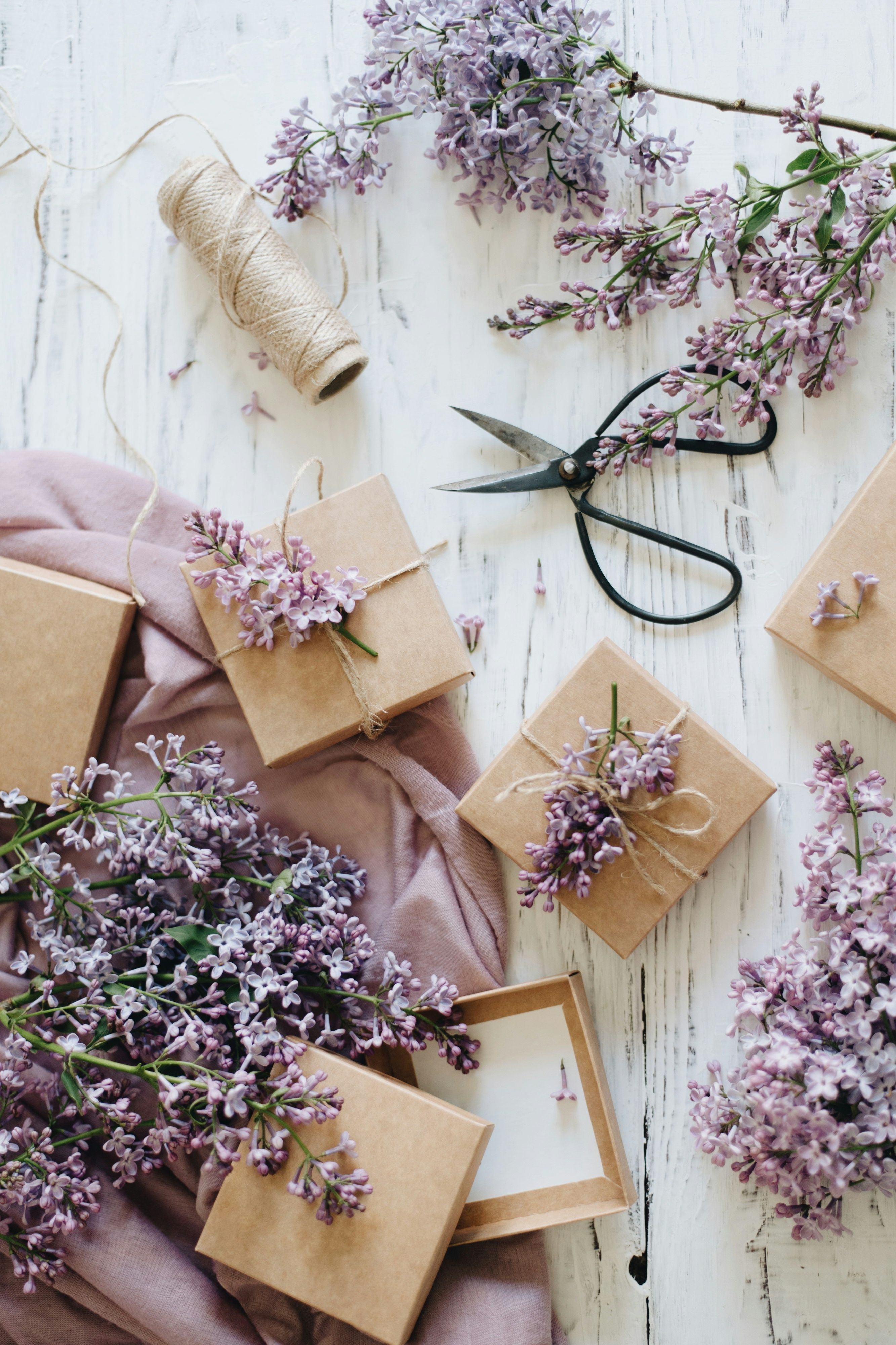 Brown gift boxes decorated with lilac flowers and twine on a white wooden table, surrounded by lilac branches, scissors, and a spool of string.