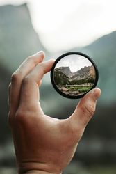 Hand holding a circular lens showing a sharp view of mountains and trees
