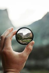 Hand holding a circular lens showing a sharp view of mountains and trees
