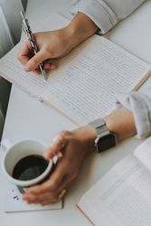Person writing in a notebook while holding a cup of coffee next to an open book.