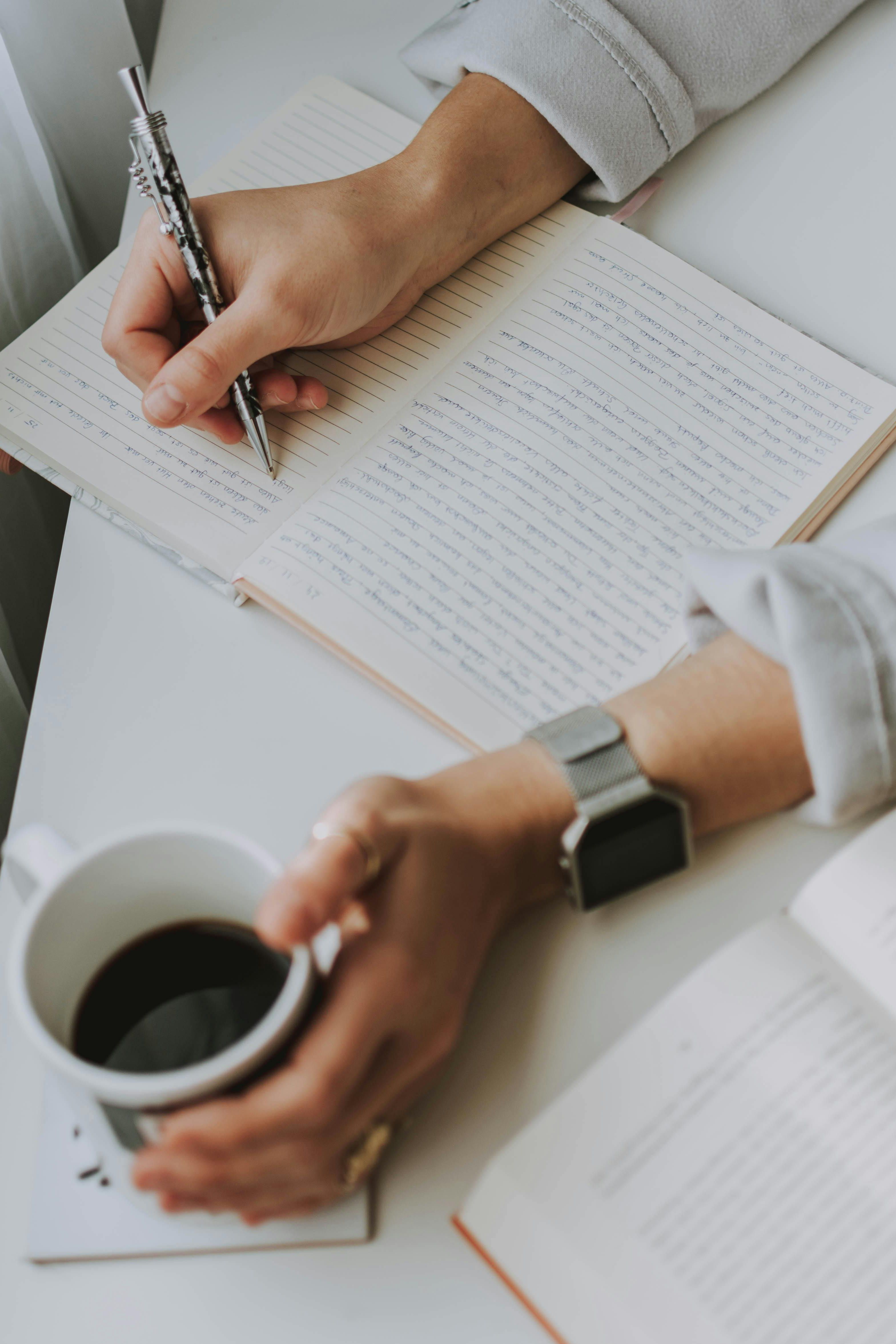 Person writing in a notebook while holding a cup of coffee next to an open book.