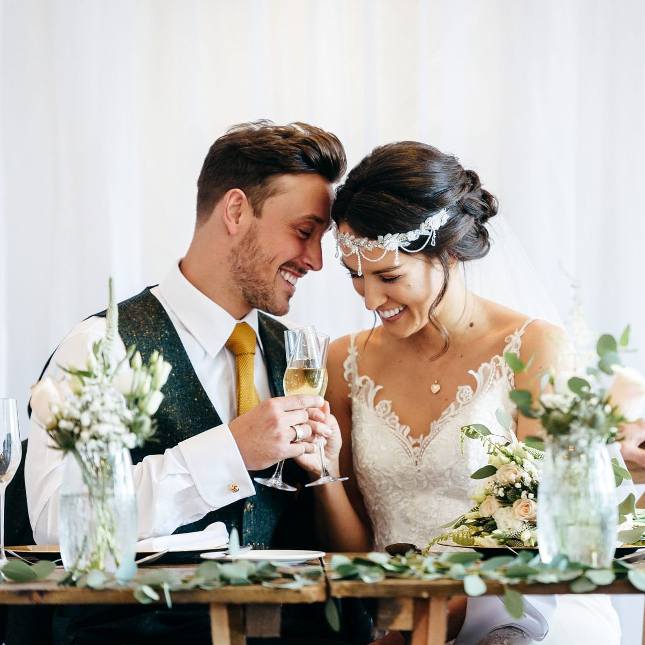 Bride and groom sitting together at a decorated table, smiling and toasting with champagne on their wedding day.