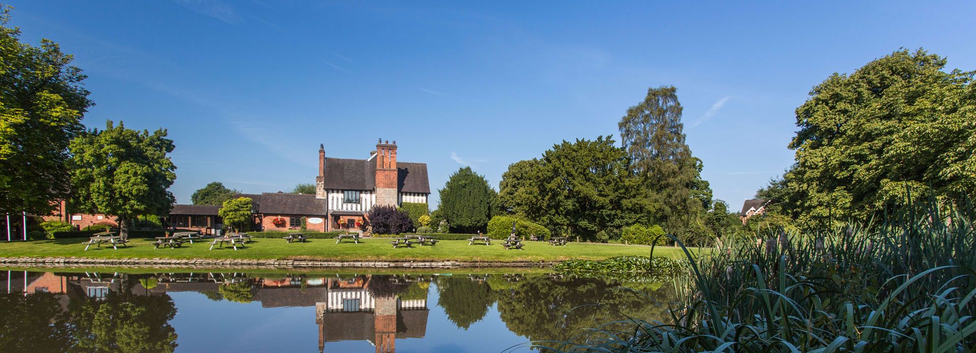 Historic building with black and white timber framing reflected in a pond, surrounded by trees and green lawns with picnic benches