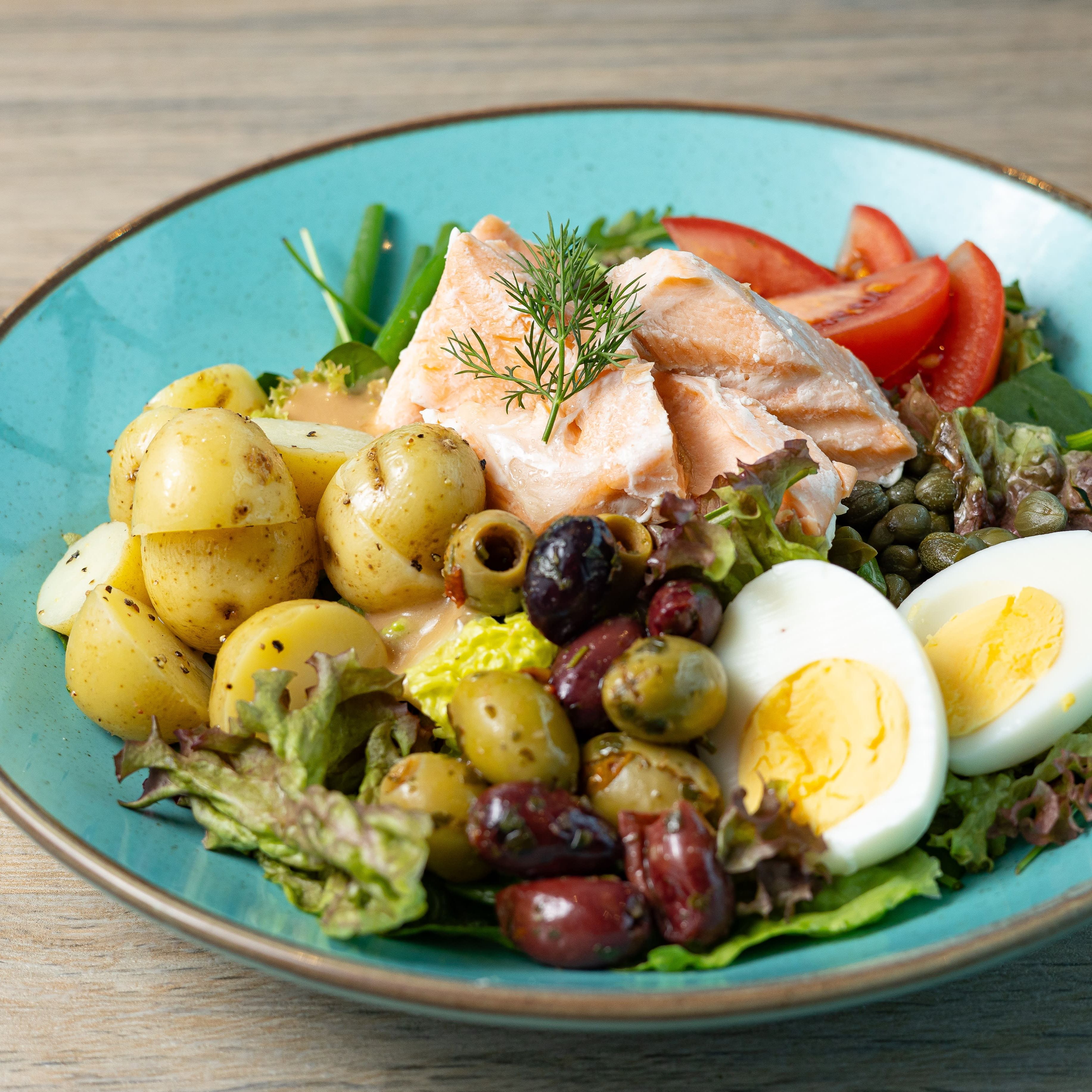 Nicoise salad with salmon, potatoes, hard-boiled eggs, tomatoes, olives, and greens on a blue plate