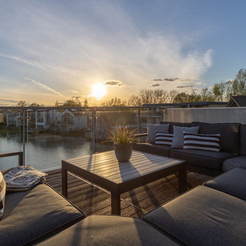 Modern outdoor patio with dark cushioned seating overlooking a lake at sunset