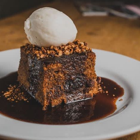 A slice of sticky toffee pudding topped with a scoop of vanilla ice cream and sprinkled with chopped nuts, served on a white plate with caramel sauce.