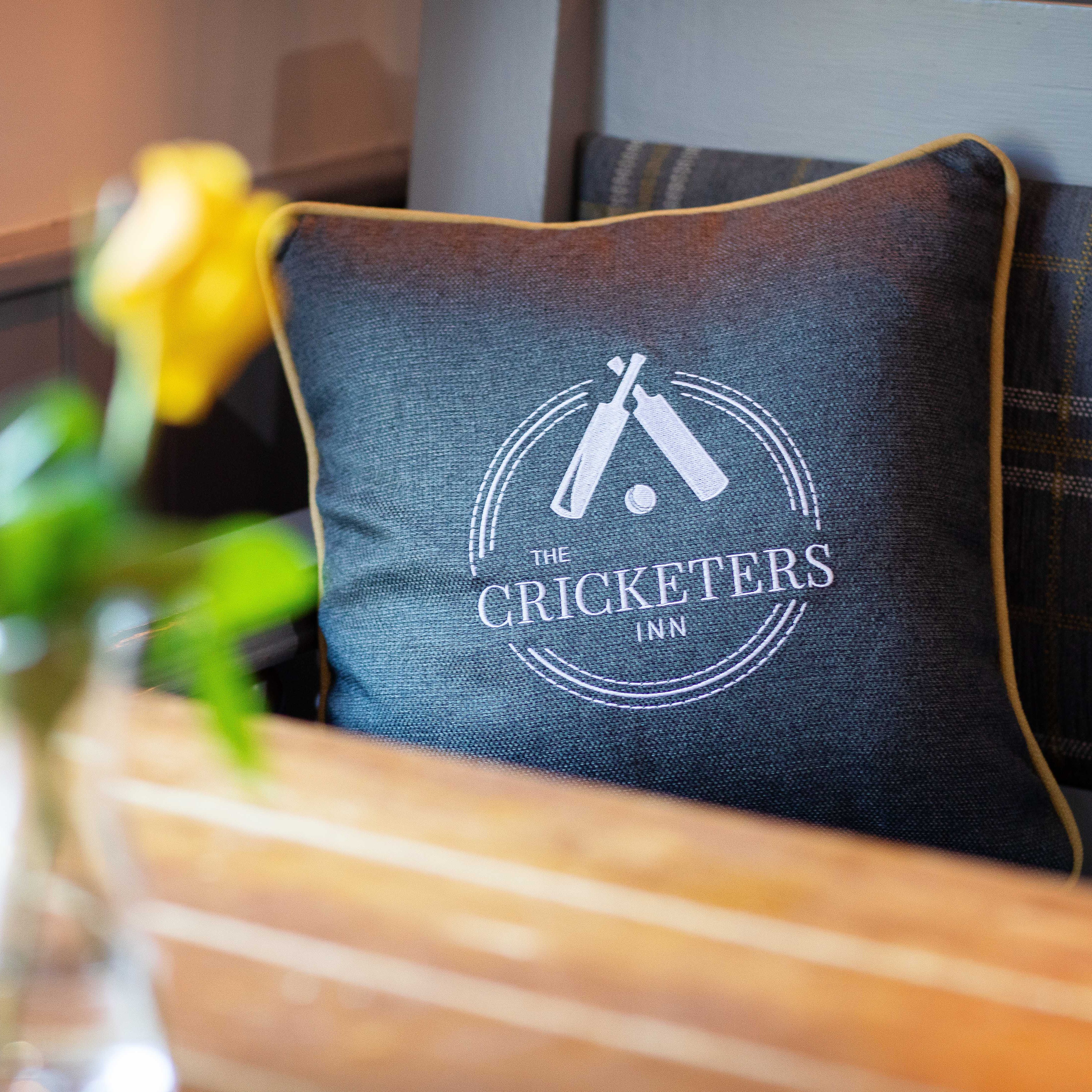 A decorative pillow with 'The Cricketers Inn' logo on a bench, next to a wooden table with yellow flowers.