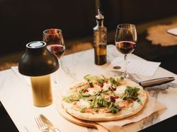 Table set with a pizza topped with arugula, two glasses of red wine, a bottle of olive oil, and a lamp