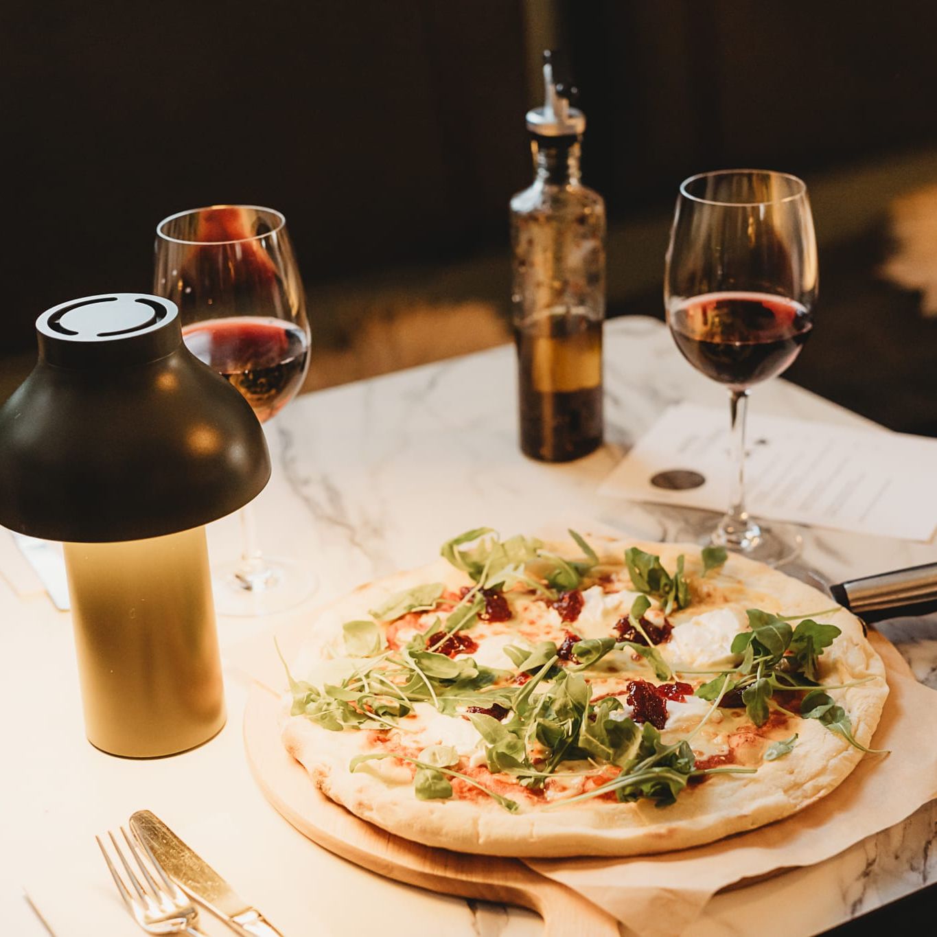 Table set with a pizza topped with arugula, two glasses of red wine, a bottle of olive oil, and a lamp