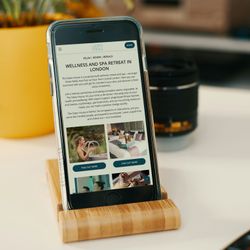 Smartphone displaying a wellness and spa retreat listing webpage in London, placed on a wooden stand next to a yellow plant pot.